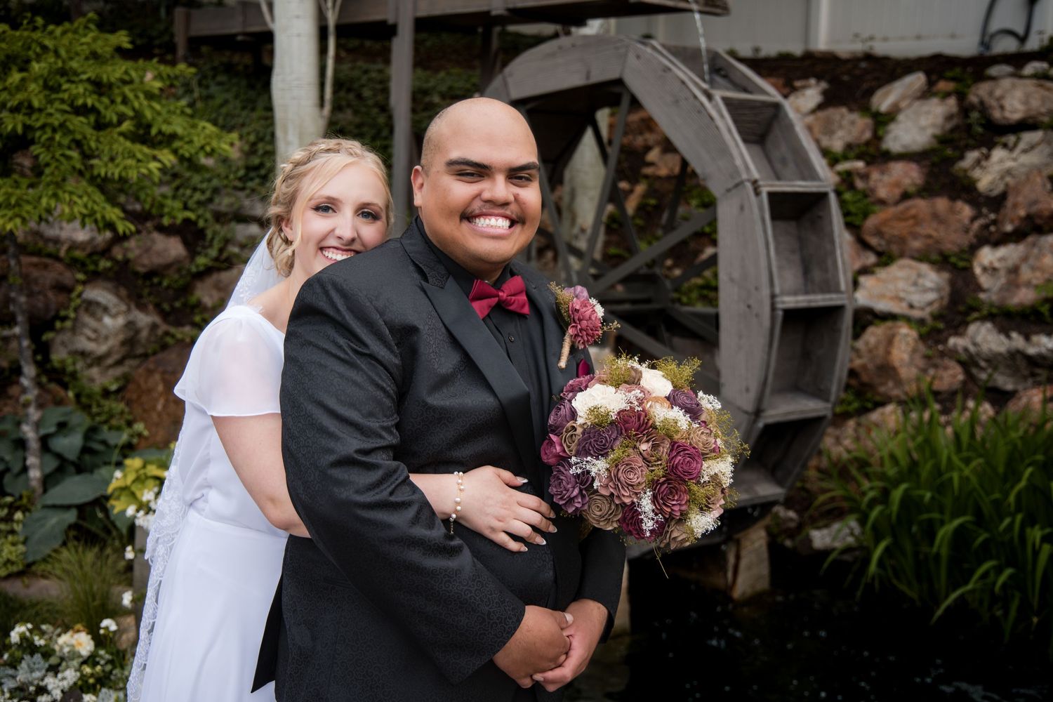 A joyful wedding couple embraces outdoors during their special day near a rustic stone wall and garden setting.