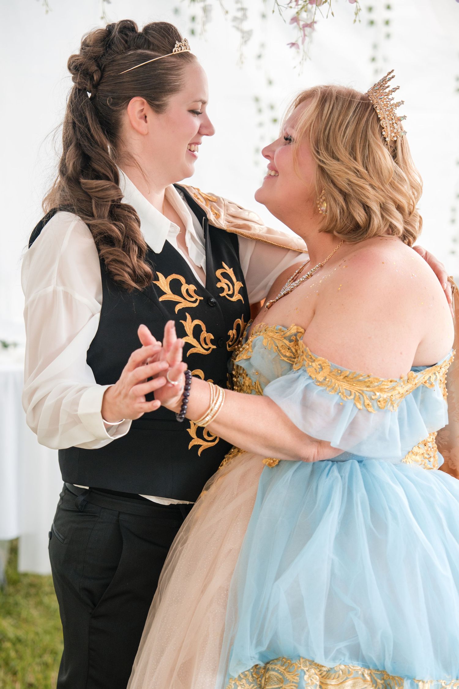 Two brides in fairy tale-inspired costumes dance together, one in a vest and one in a flowing blue gown with gold details.