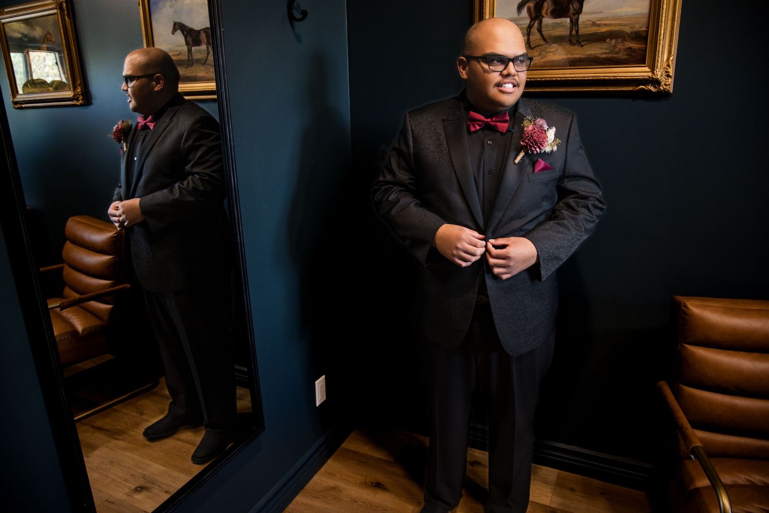 A man in a formal black suit stands against dark blue walls in an elegant interior setting.
