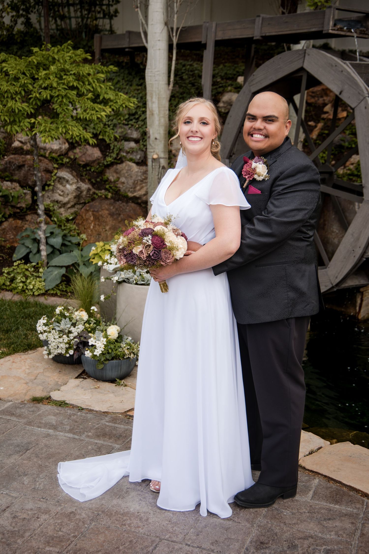 A newly married couple poses by a rustic water wheel at an outdoor garden wedding venue with floral decorations.
