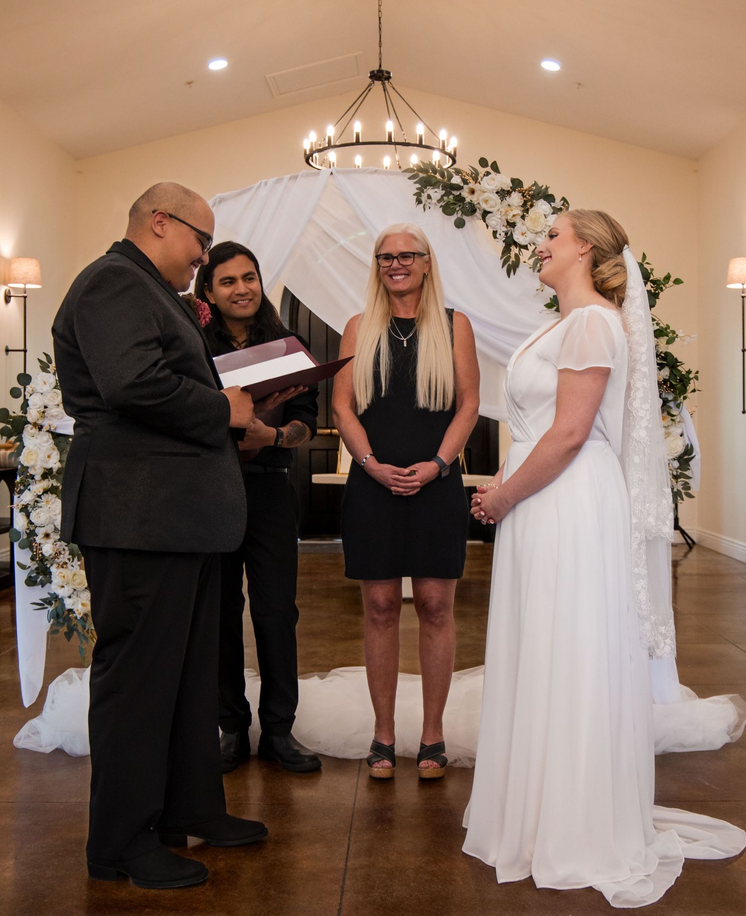 A wedding ceremony takes place inside a chapel with a white floral arch and elegant chandelier lighting.