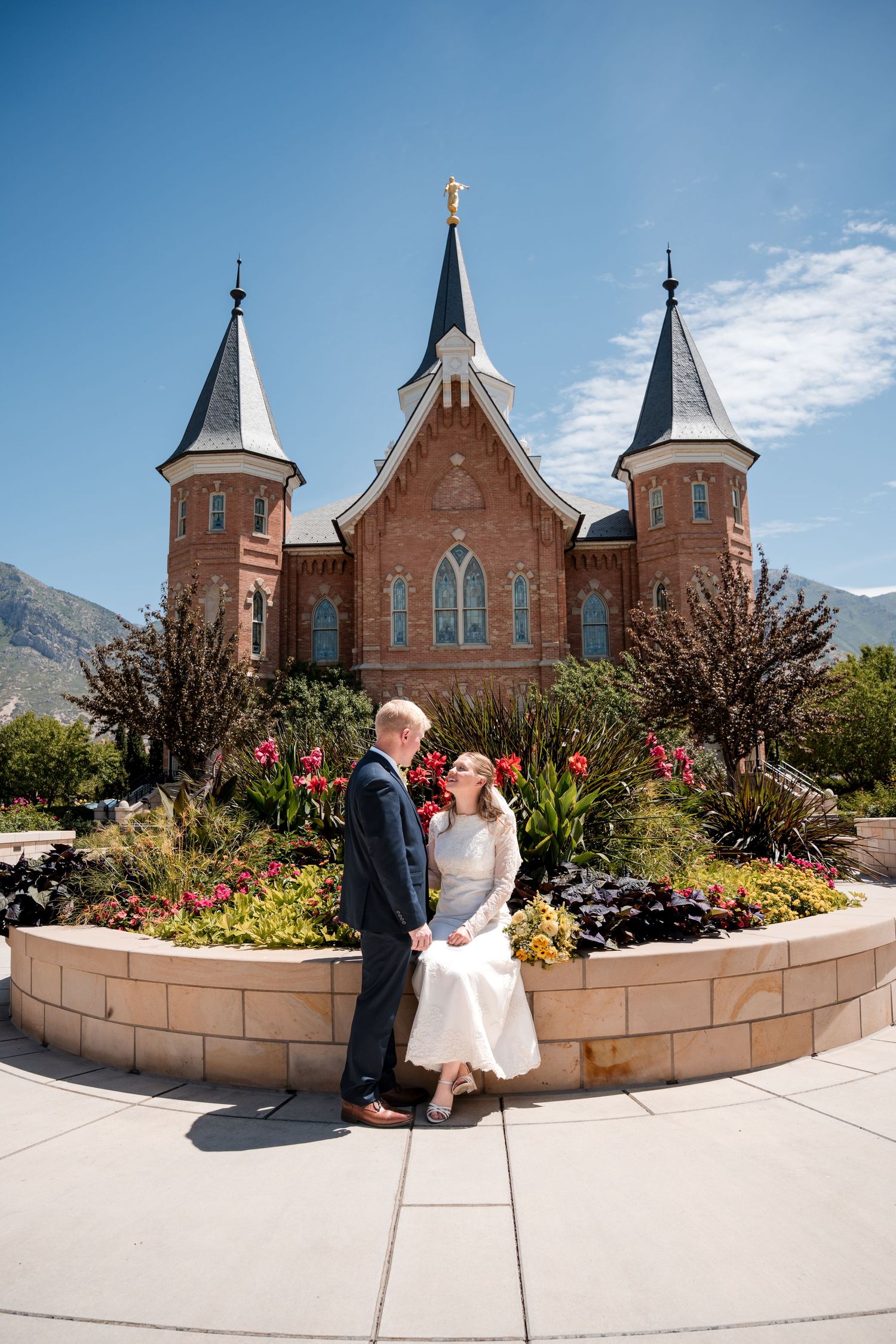 A couple poses in formal attire outside the Provo City Center LDS temple with three spires against a blue sky and mountain backdrop.