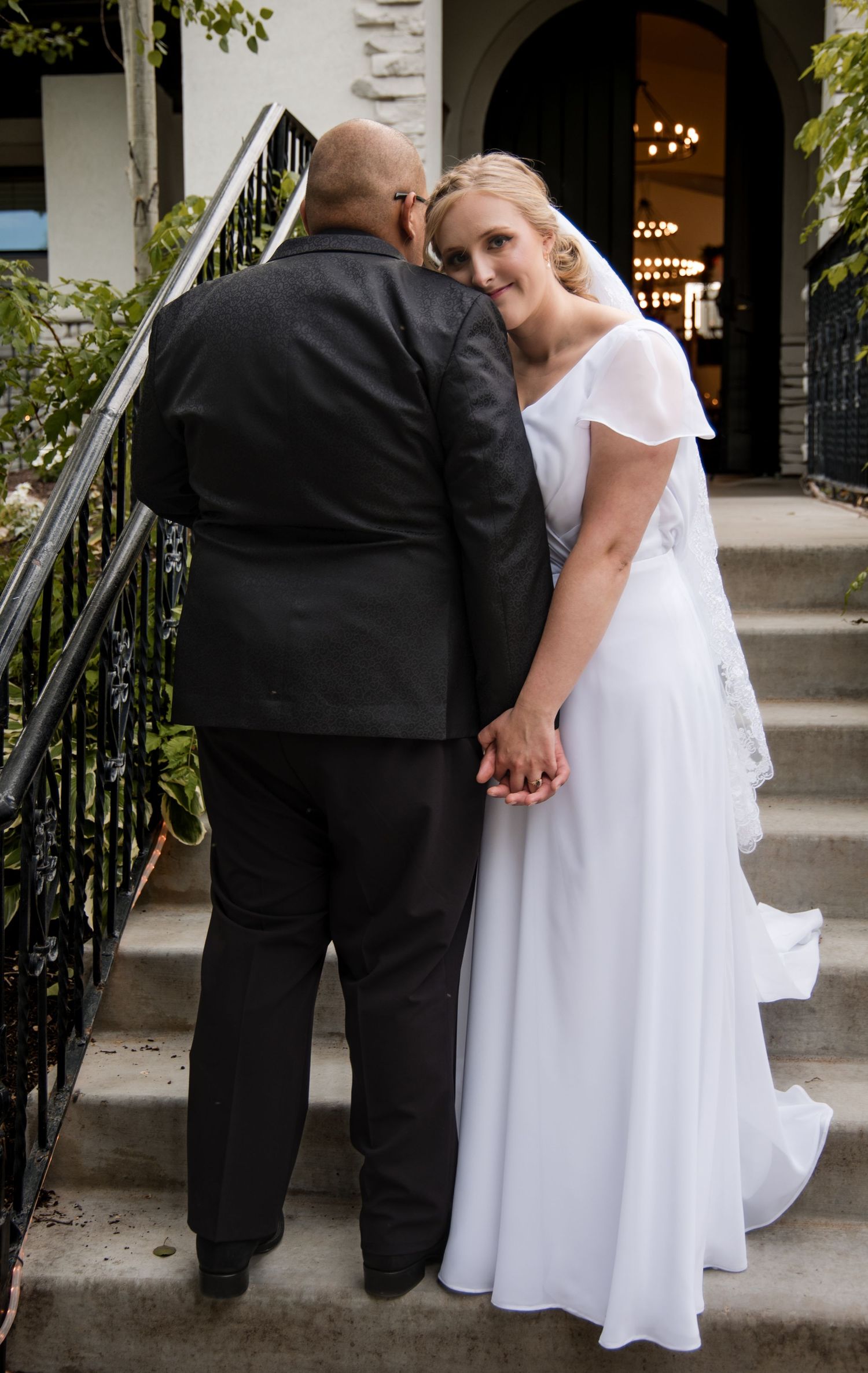 A couple shares an intimate moment on outdoor steps, with the person in white wearing an elegant wedding dress.