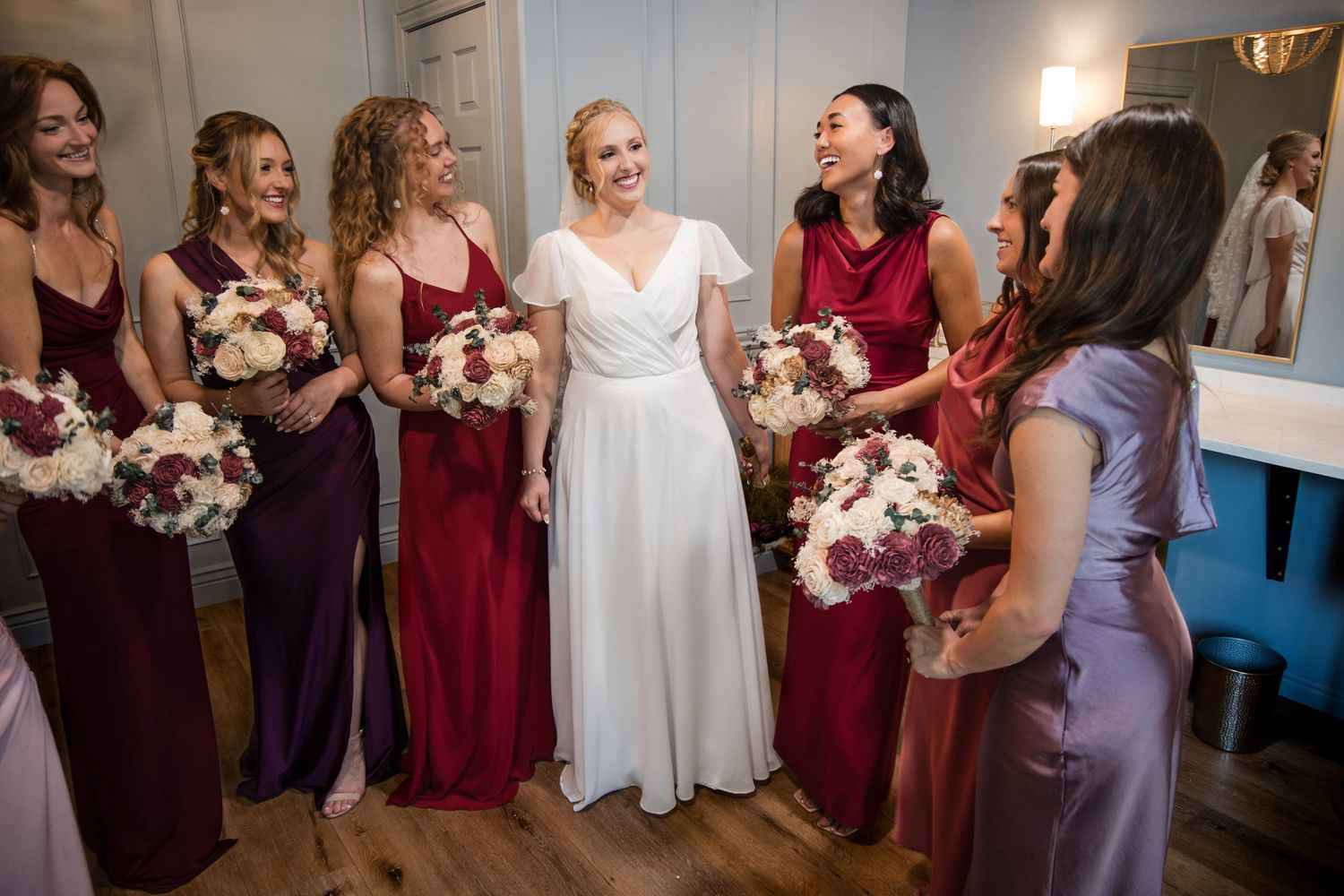 Wedding party in burgundy and purple bridesmaids dresses gather together holding floral bouquets during a wedding.