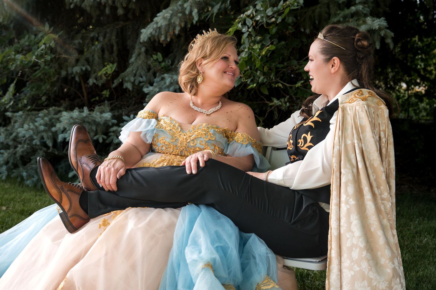 Two people in princess costume dresses relax and laugh together on a bench in an outdoor garden setting.