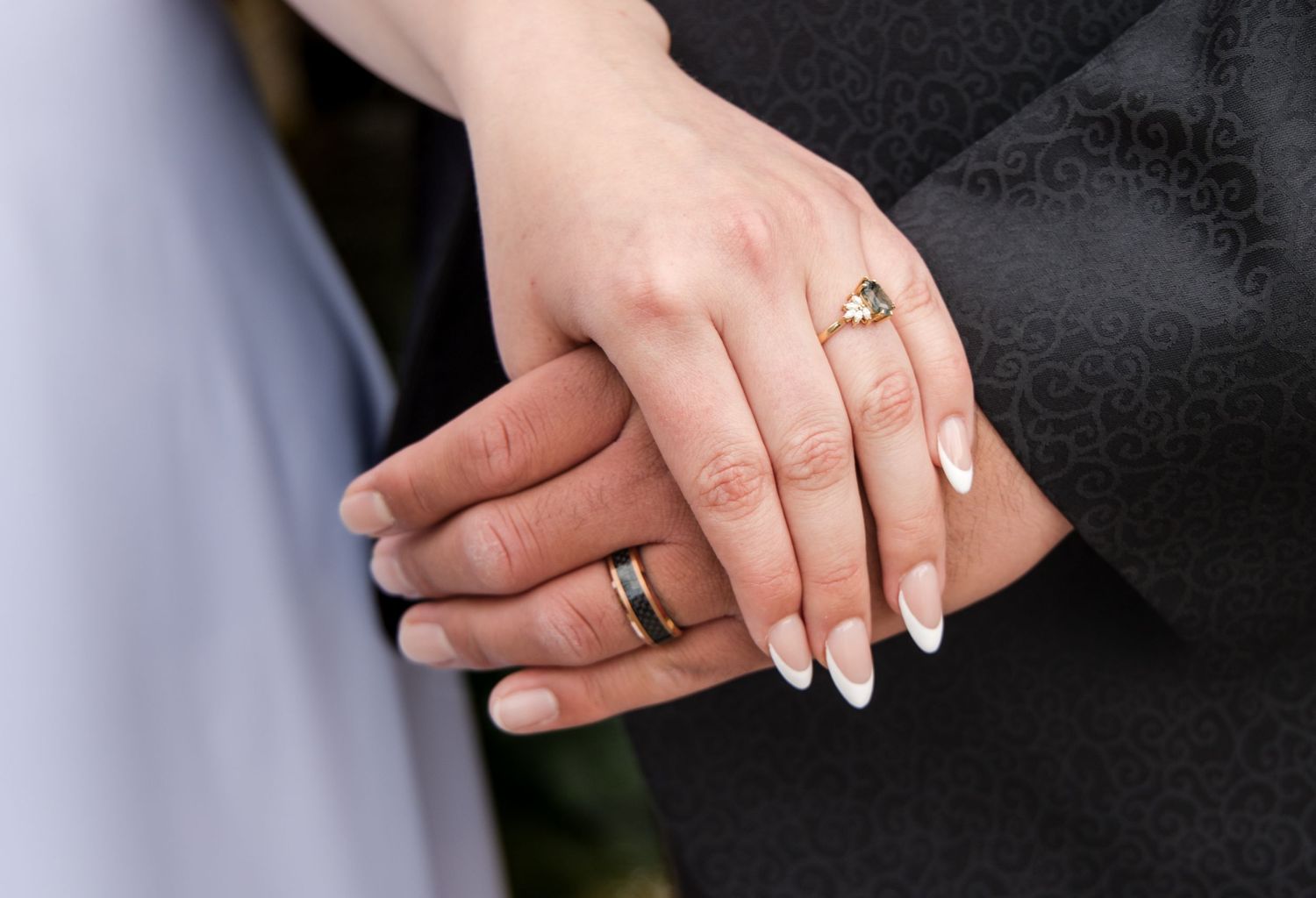 Close-up photo of newlywed couple's hands showing wedding rings and white manicured nails against black and white attire.