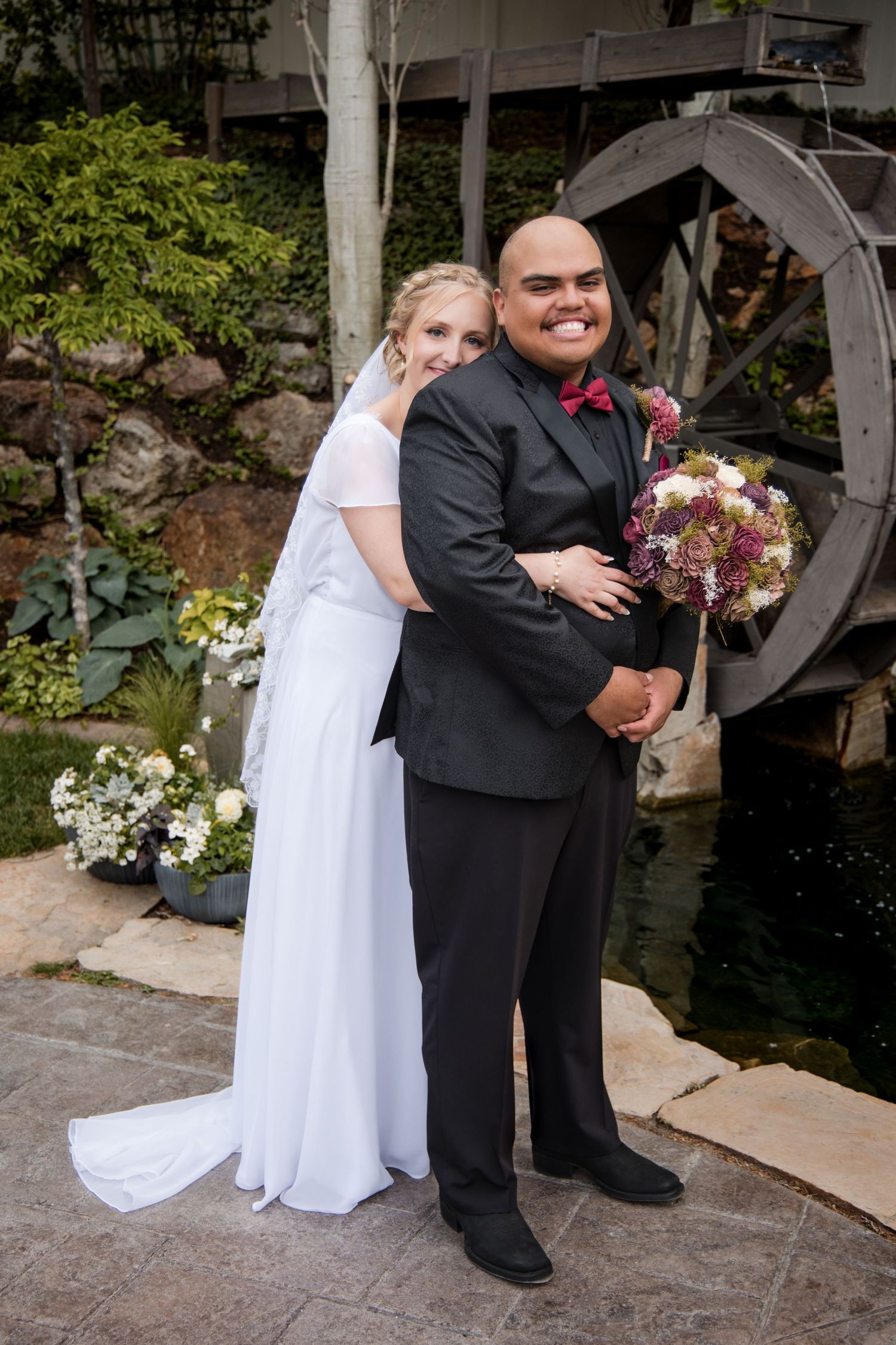 A couple poses in formal wedding attire in front of a garden water feature with flowers and greenery.