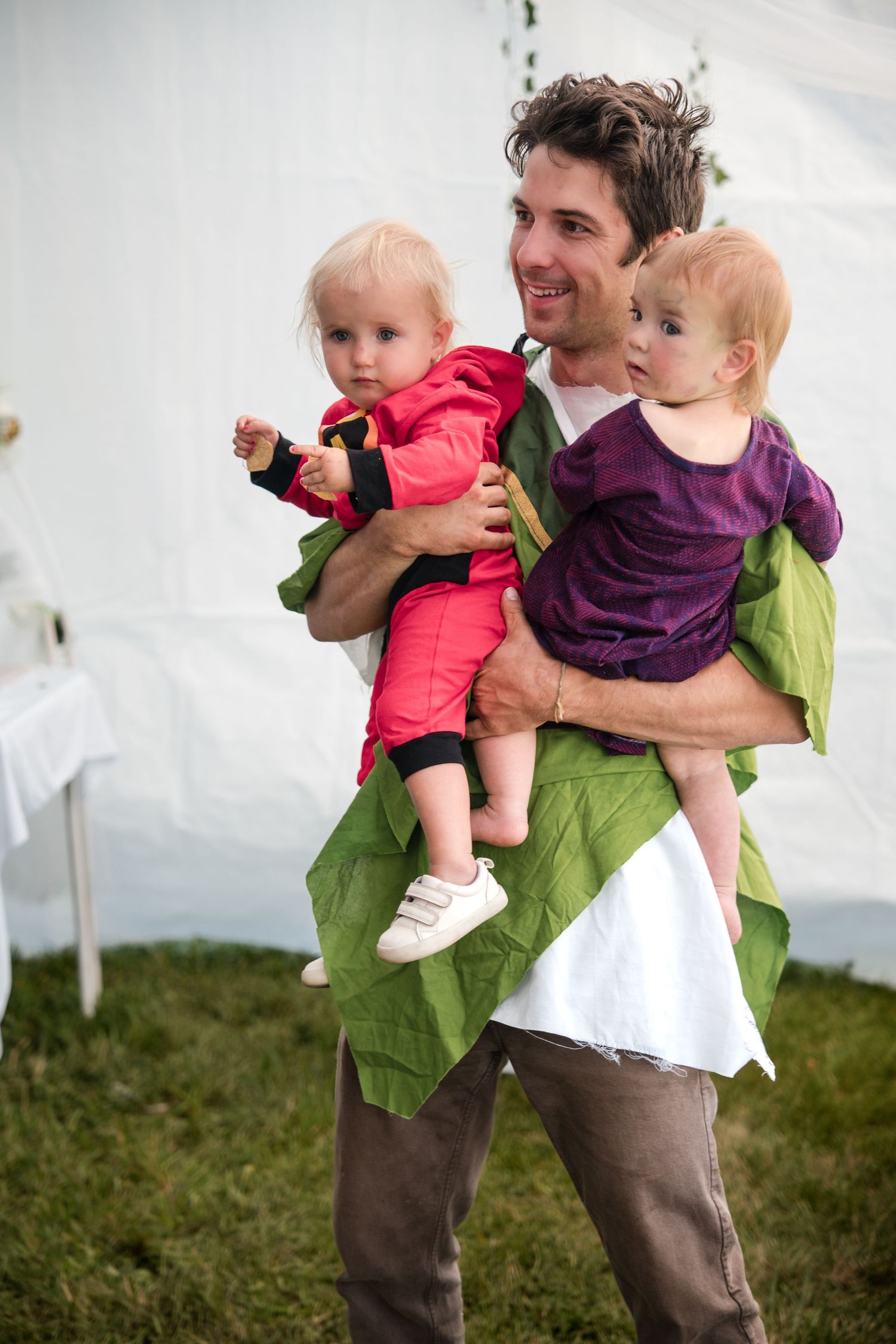 Parent in green shirt holds two small children while standing on grassy hill with white tent in background.