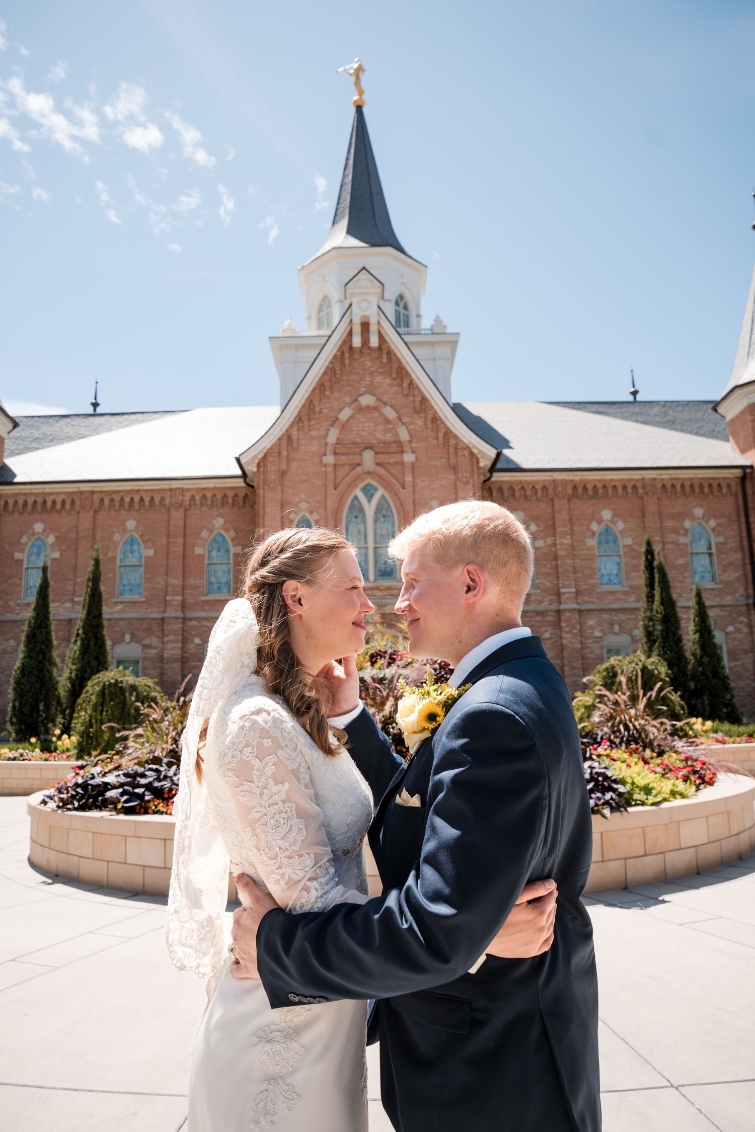 Just married couple embraces affectionately outside a Mormon temple surrounded by lush greenery and spires.