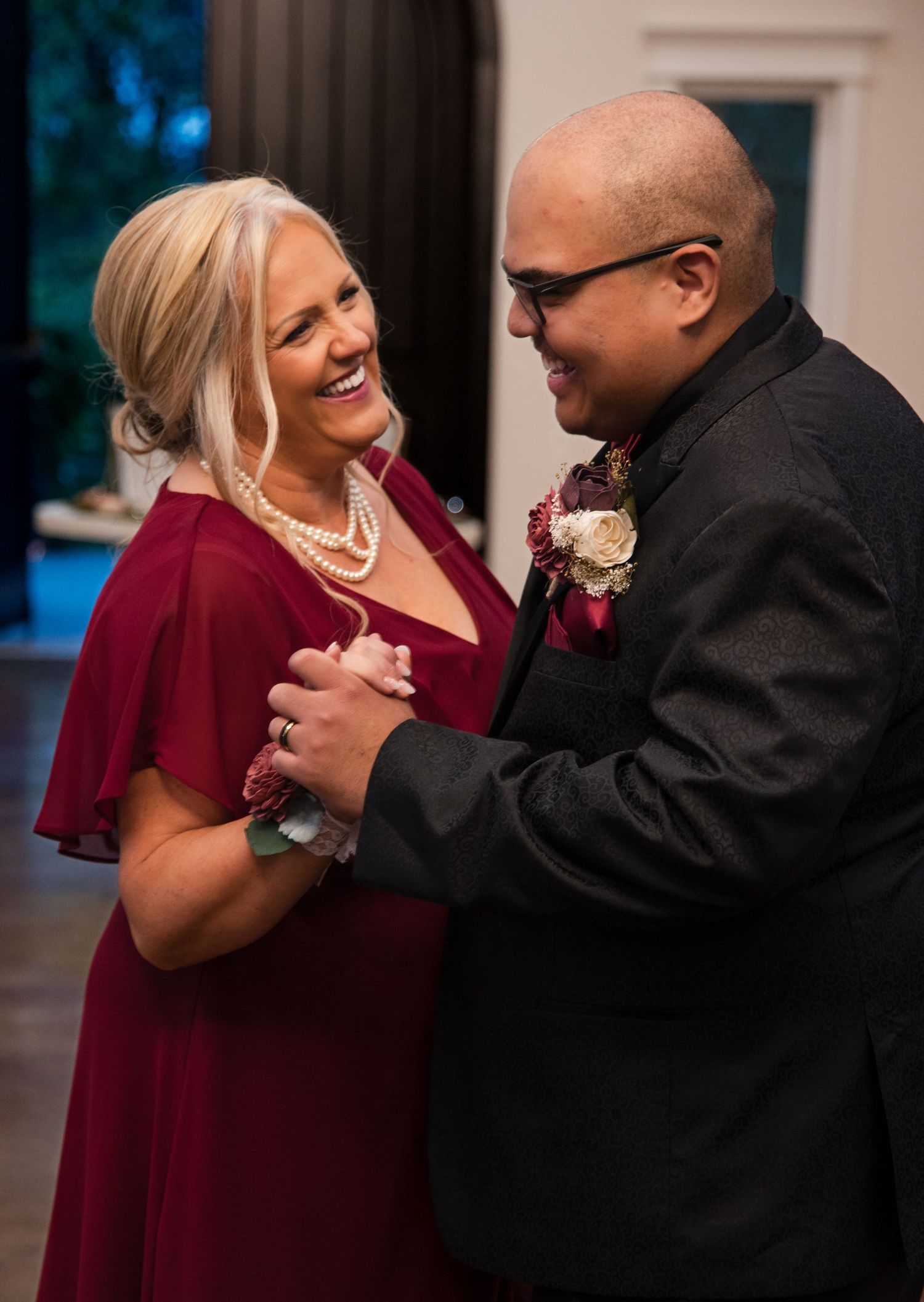 A couple smiles while sharing an intimate dance at a formal event, with the woman in burgundy and man in black attire.
