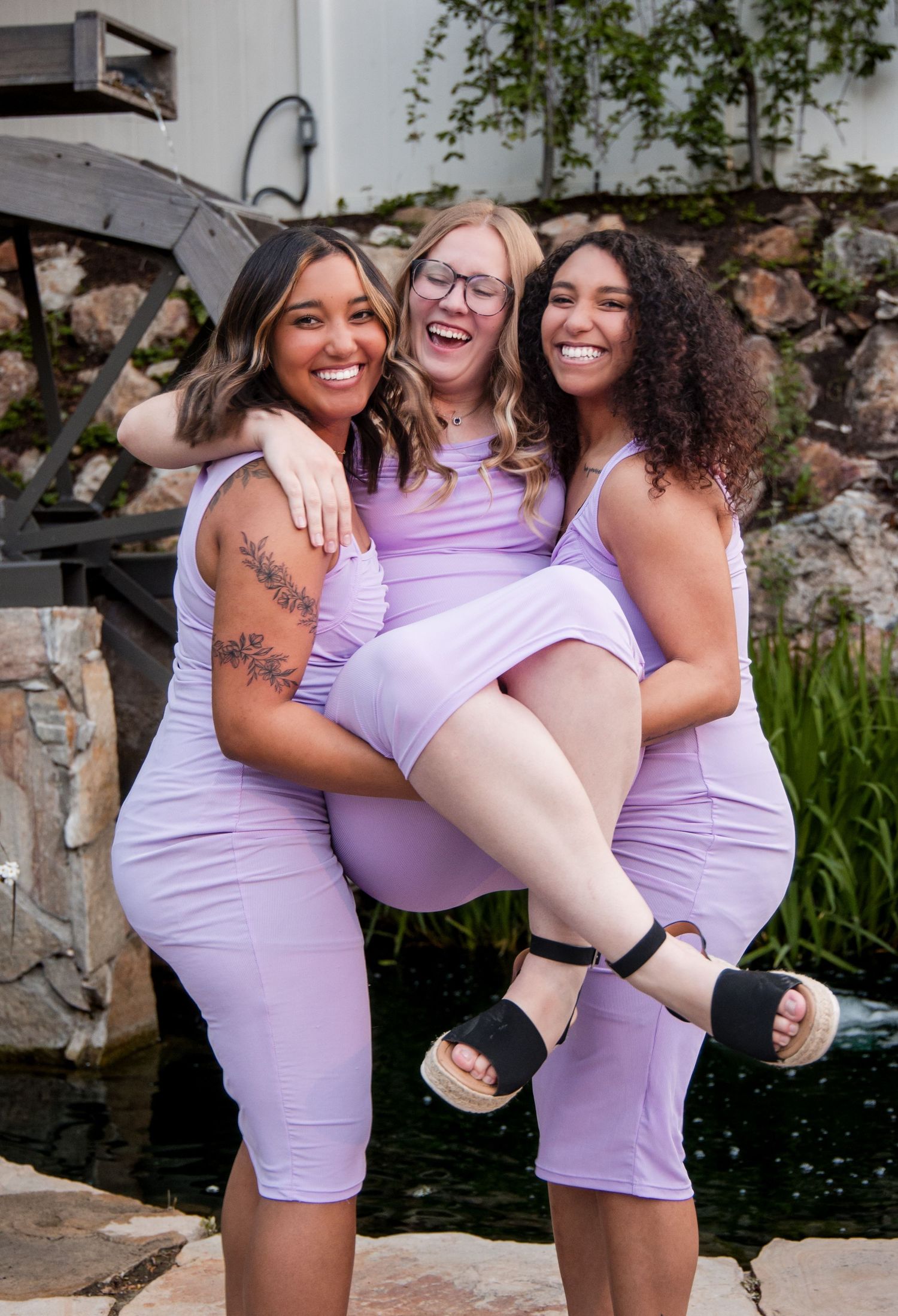 Group wearing matching purple dresses poses playfully together for photos outdoors near stone wall.