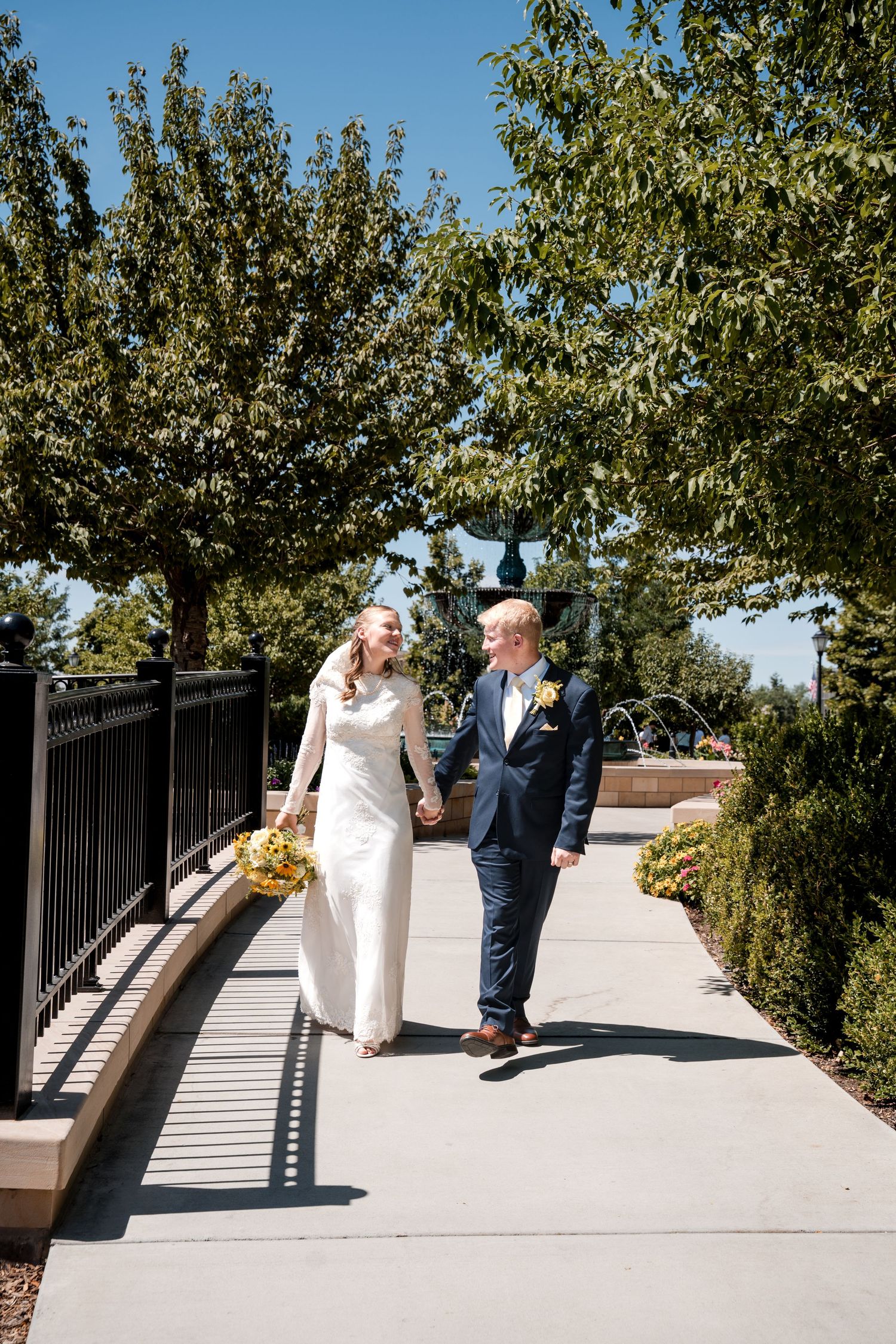 A couple in wedding attire walks together along a sunny outdoor walkway lined with trees on a beautiful summer day.