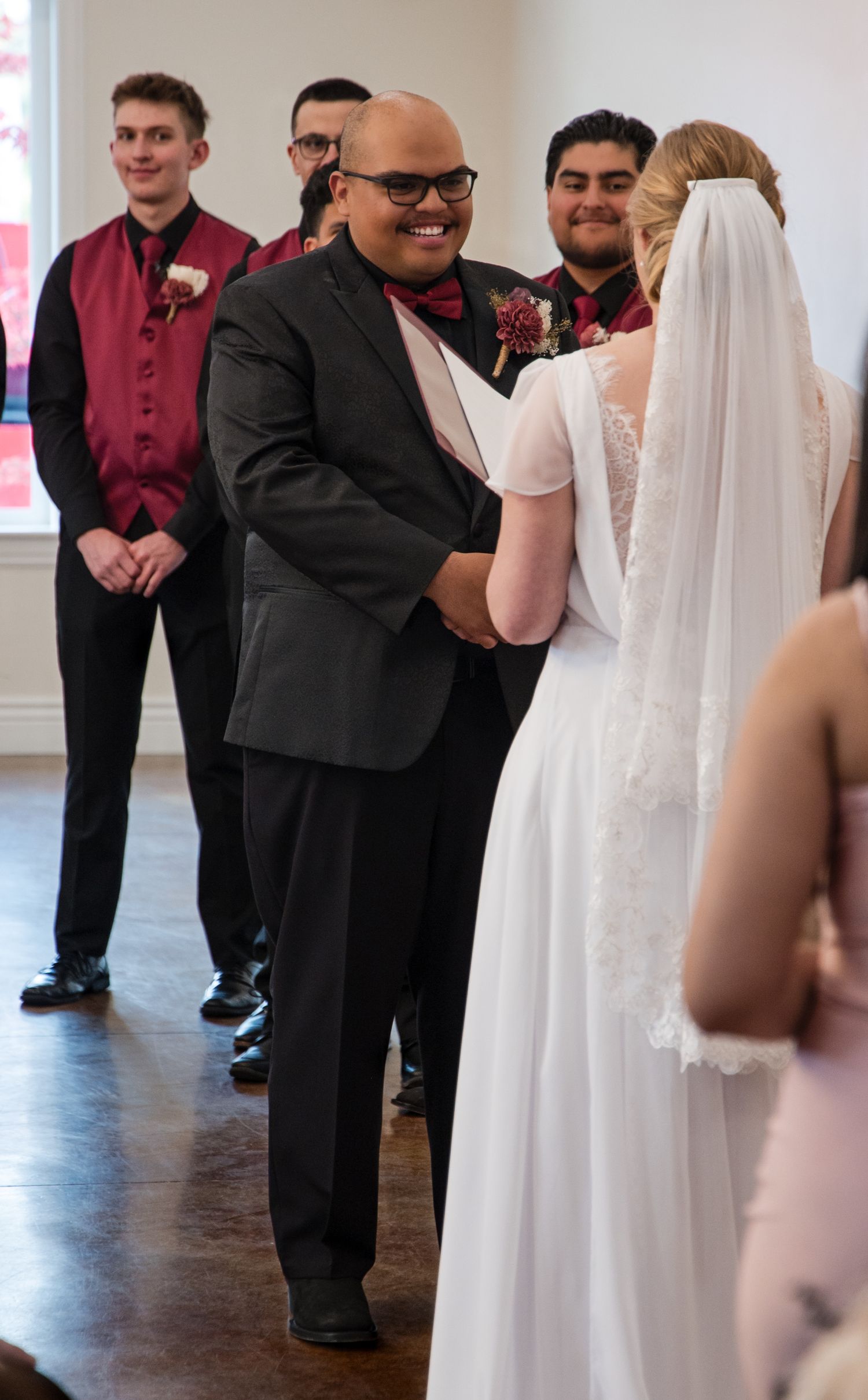 Wedding party members in burgundy and black attire stand in line during ceremony at indoor venue.
