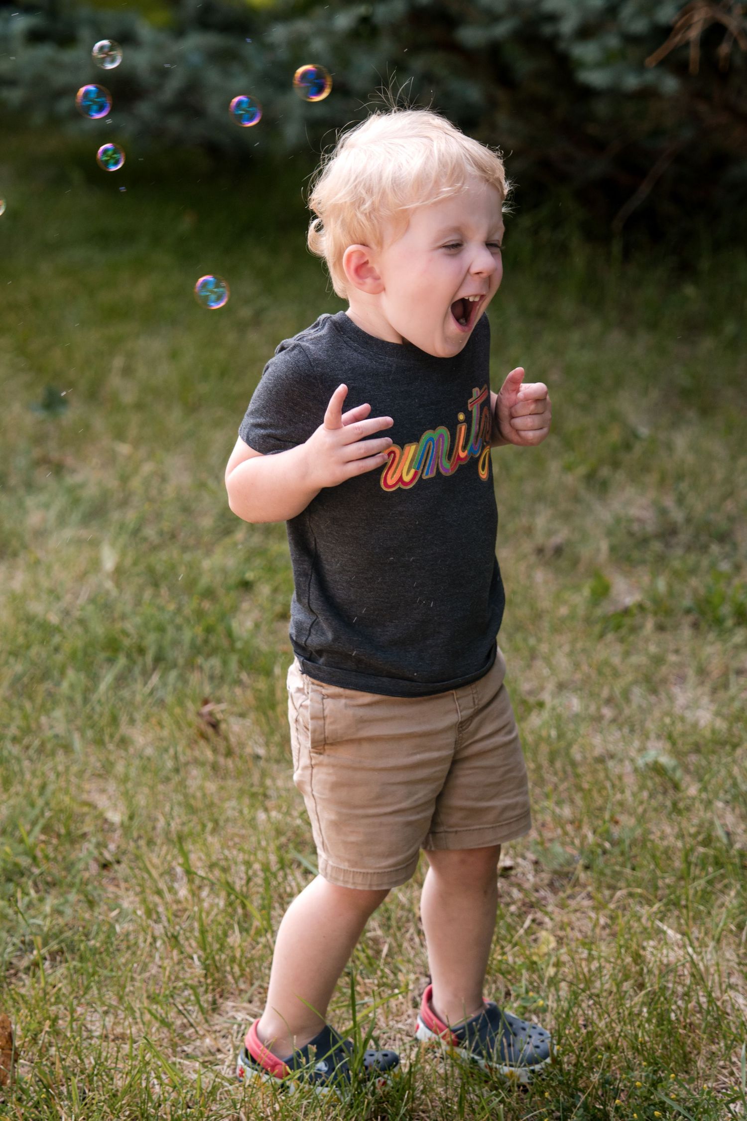 A small child in khaki shorts and dark shirt chasing bubbles with delight in a grassy yard.