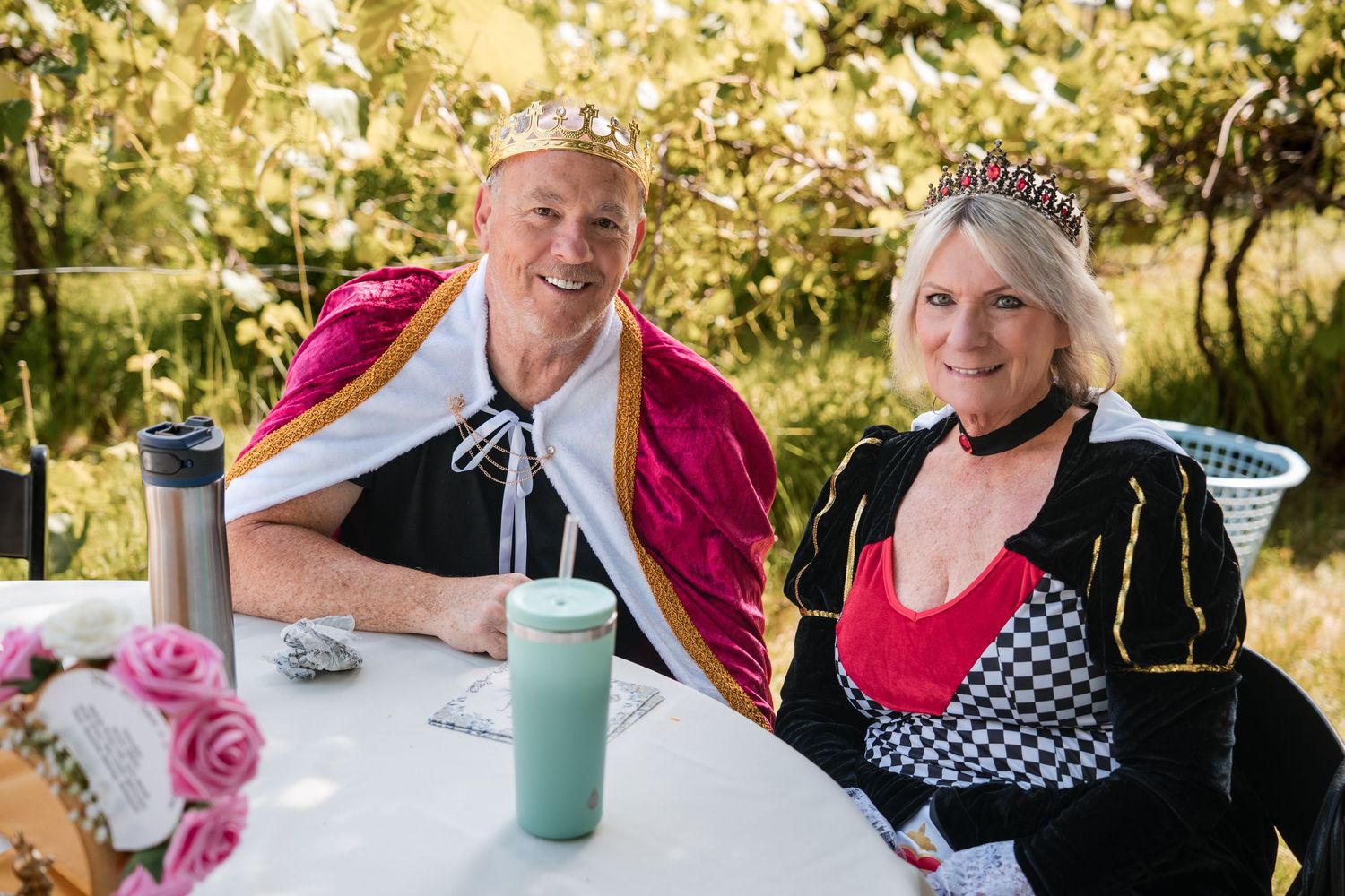 Two people in medieval royal costumes sit at an outdoor table with roses and beverages in a sunlit garden setting.