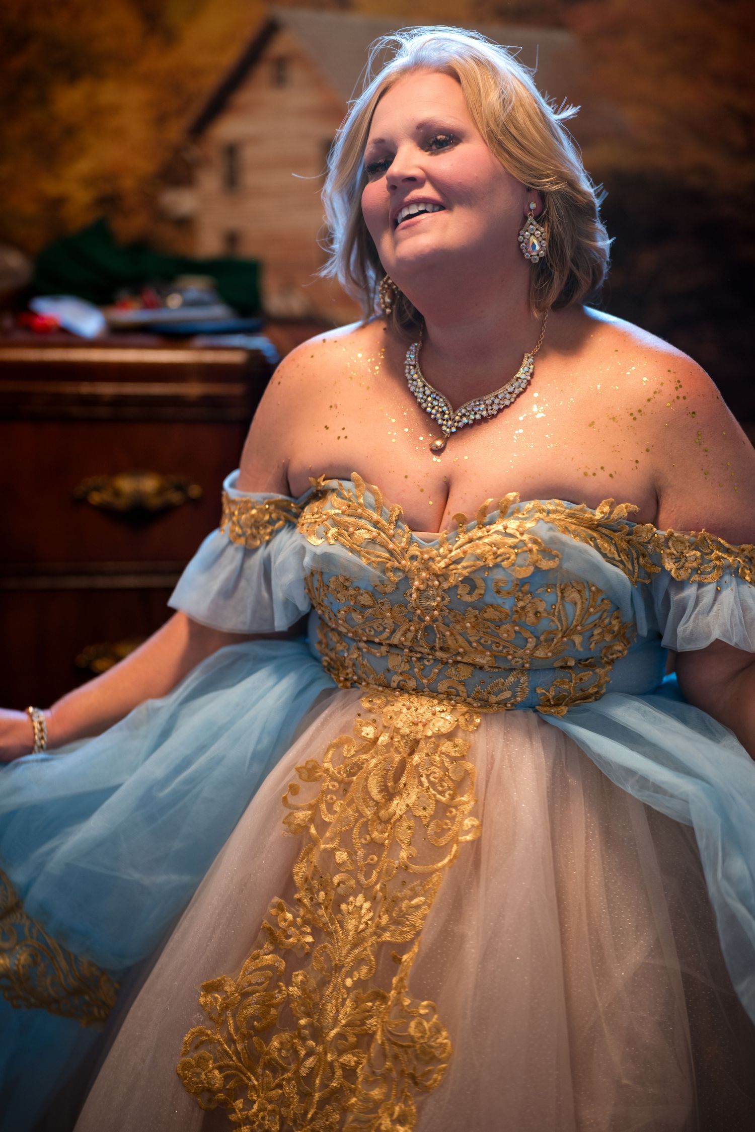 A woman wearing an elegant blue and gold ballgown with sparkles and a necklace smiles while sitting indoors.