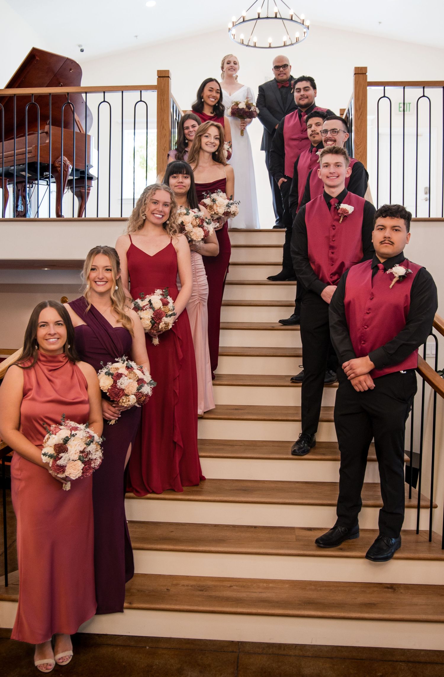 Wedding party poses on white stairs, wearing burgundy and maroon formal attire with blush pink floral bouquets.