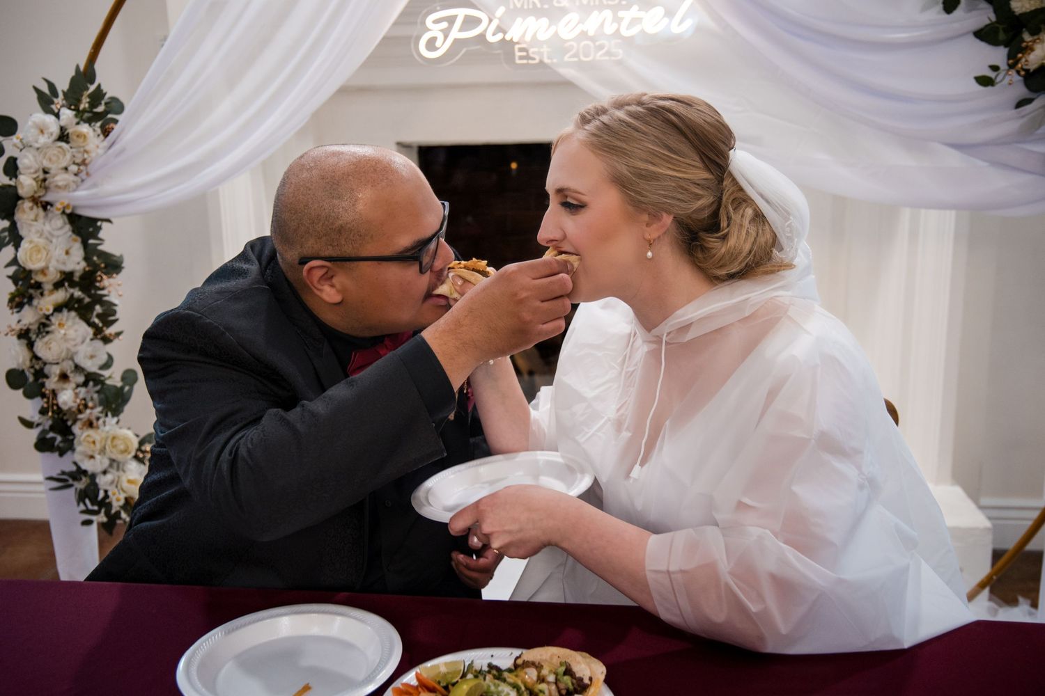 A tender wedding moment captured during dinner as a couple shares a loving bite of food under an elegant white draped canopy.