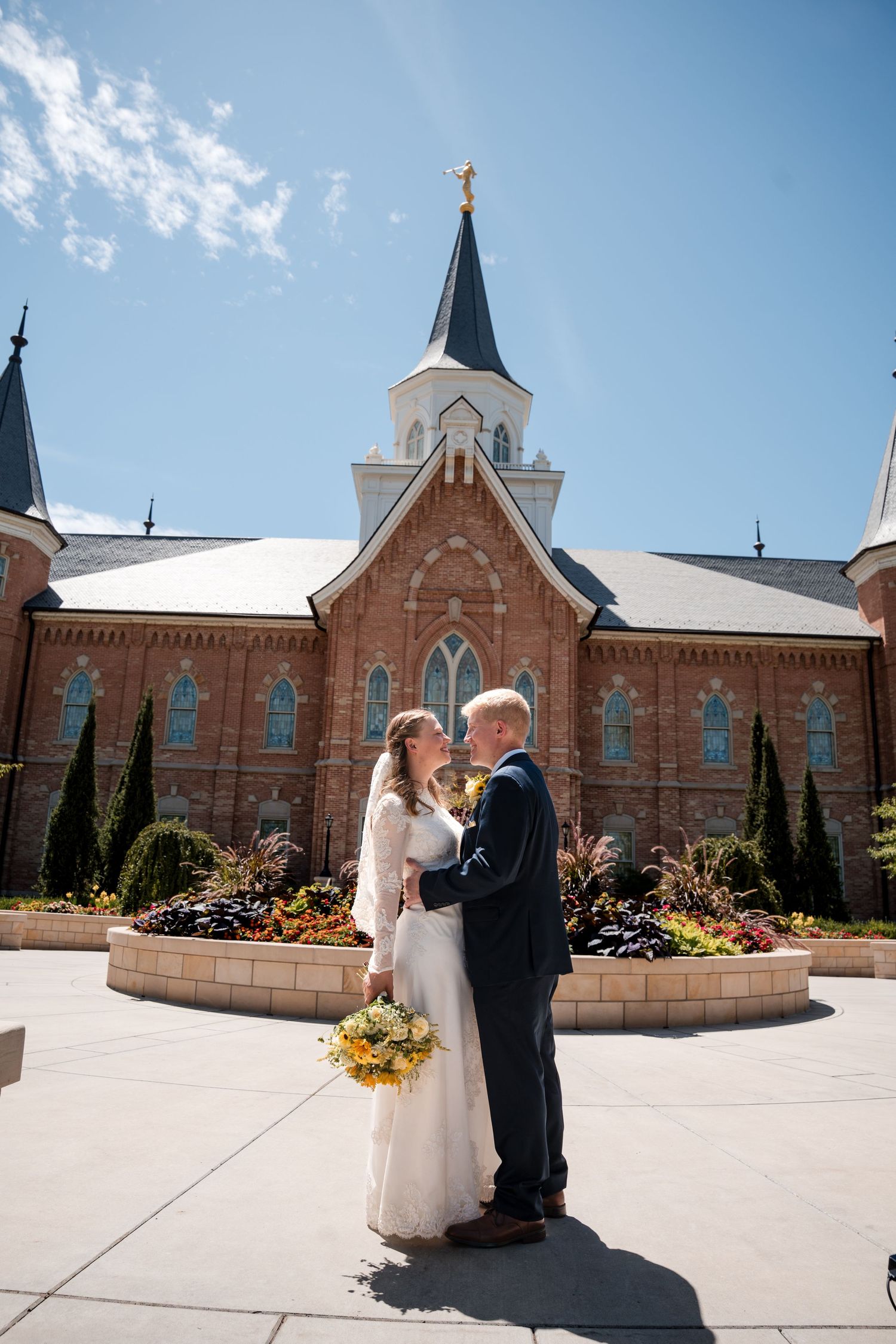 A couple shares an intimate moment in front of the Provo City Center LDS temple with spires under a bright blue sky.