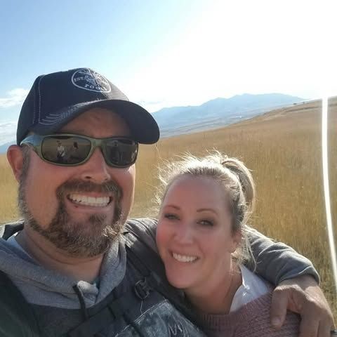 Two people taking a selfie outdoors with mountains and golden hills in the background on a sunny day.