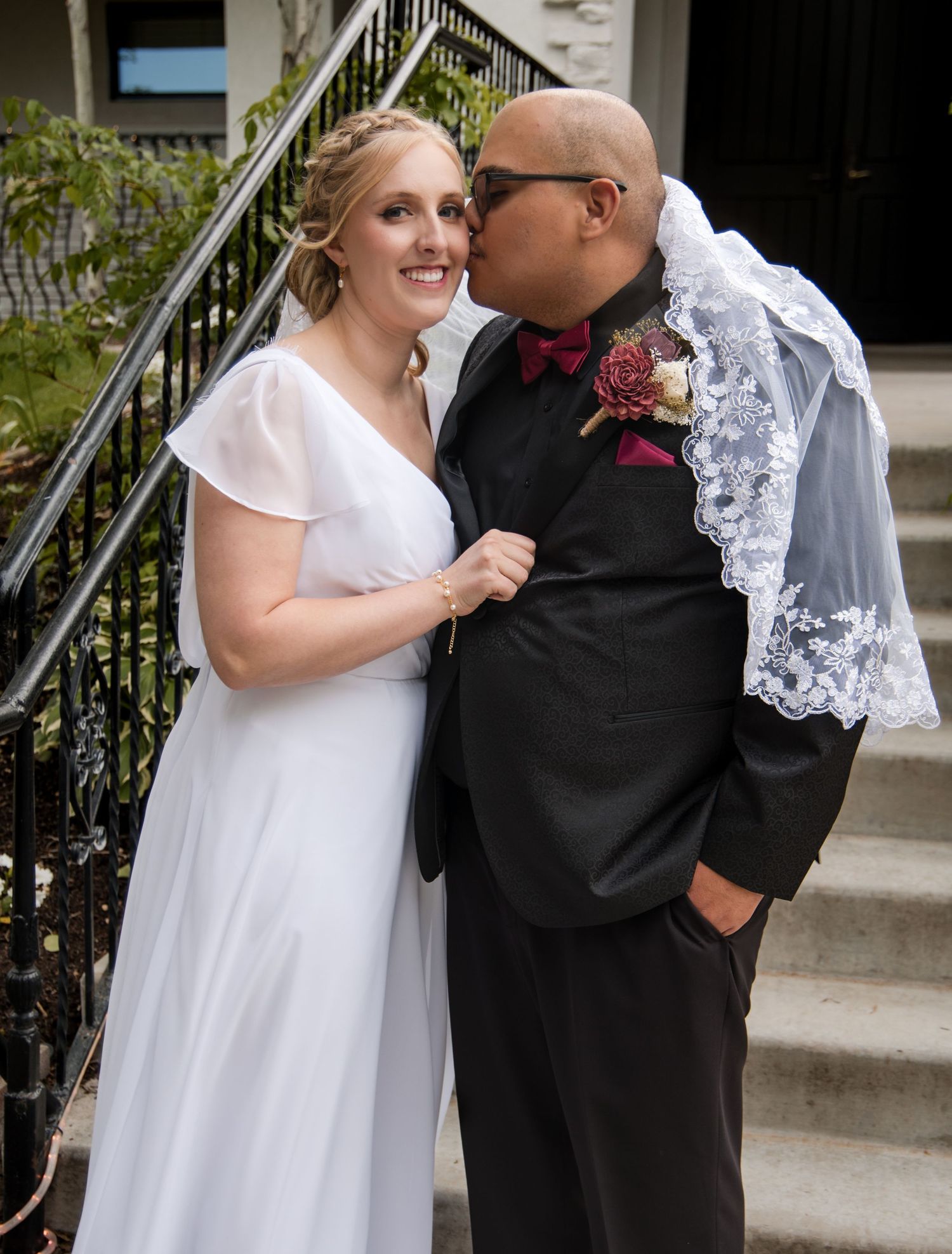 A happy newlywed couple sharing a sweet moment on outdoor steps, with the groom wearing a black suit and red bowtie.