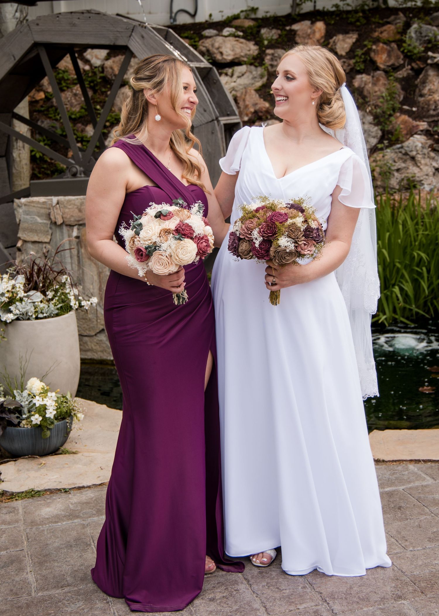Two people in purple and white dresses share joyful moments together while holding floral bouquets at an outdoor venue.