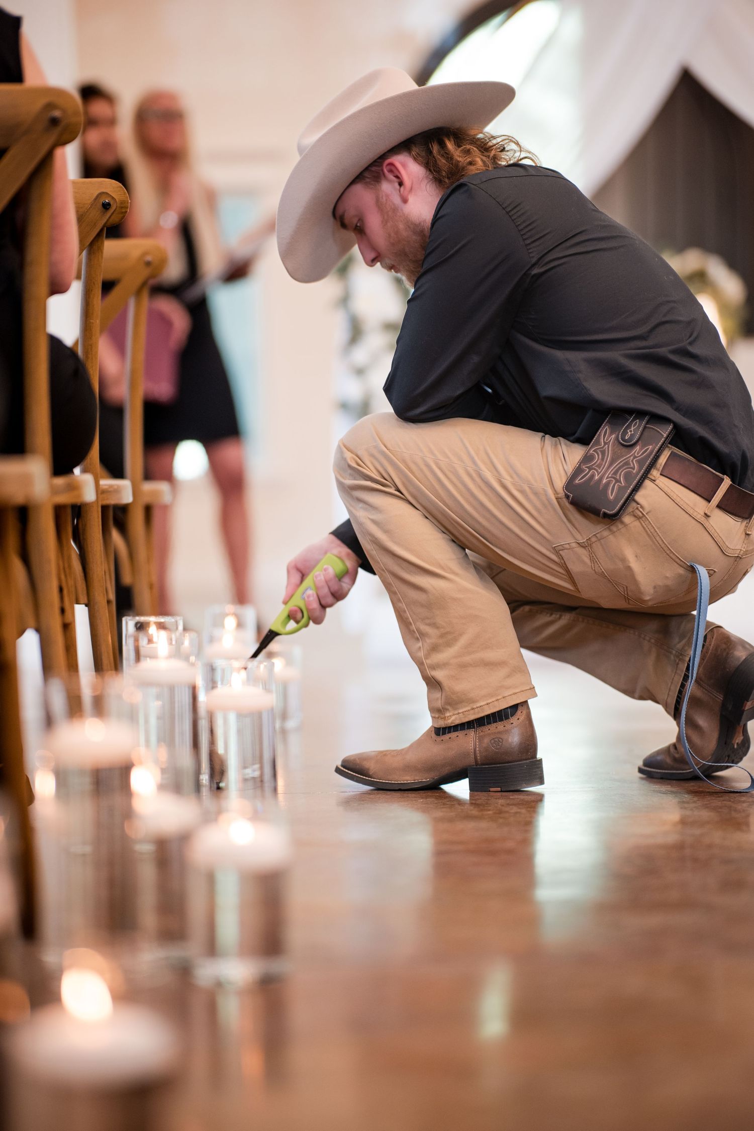 A man in cowboy boots and hat crouches down on a wooden floor to light candles at an indoor event.