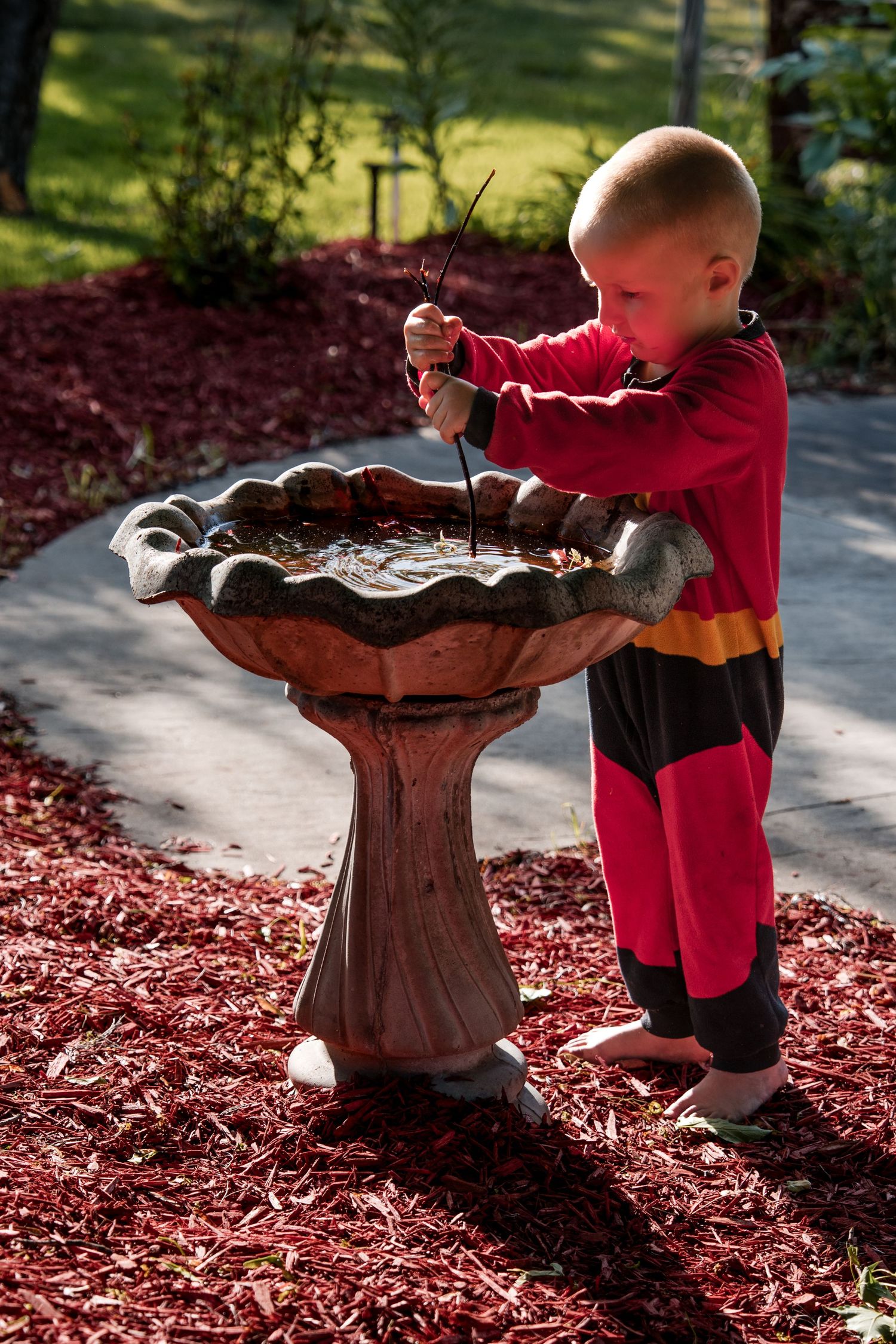 Small child wearing red outfit plays with water in decorative bird bath on mulch covered garden path.