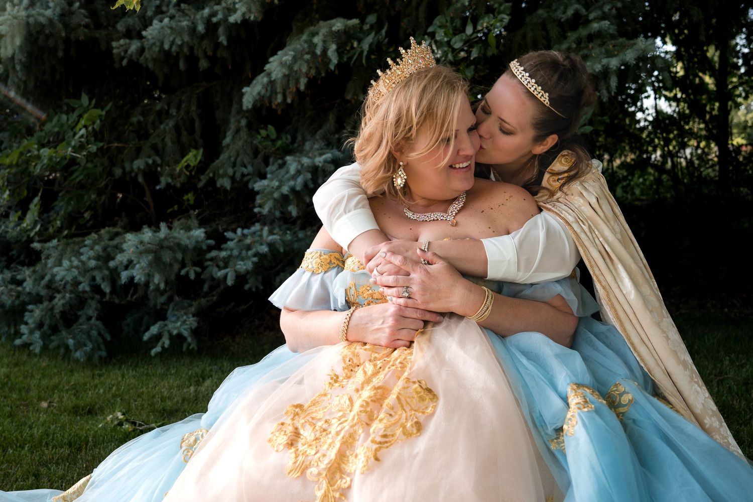 Two people in elegant period costumes with full skirts embrace while sitting together on grass with evergreen trees in background.