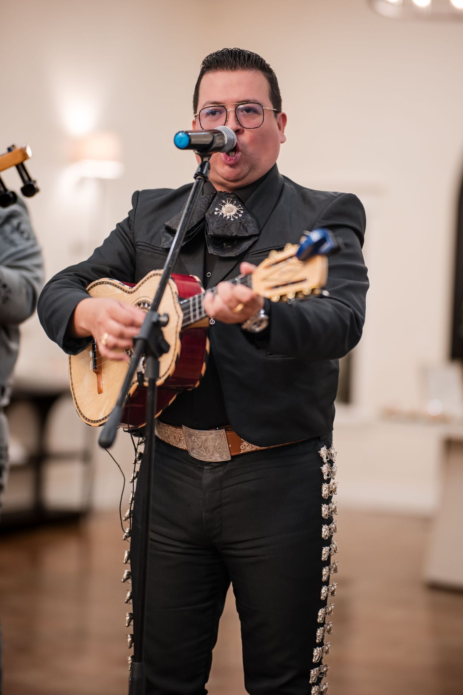 Mariachi musician in black charro suit performs with a small acoustic guitar at an indoor venue.