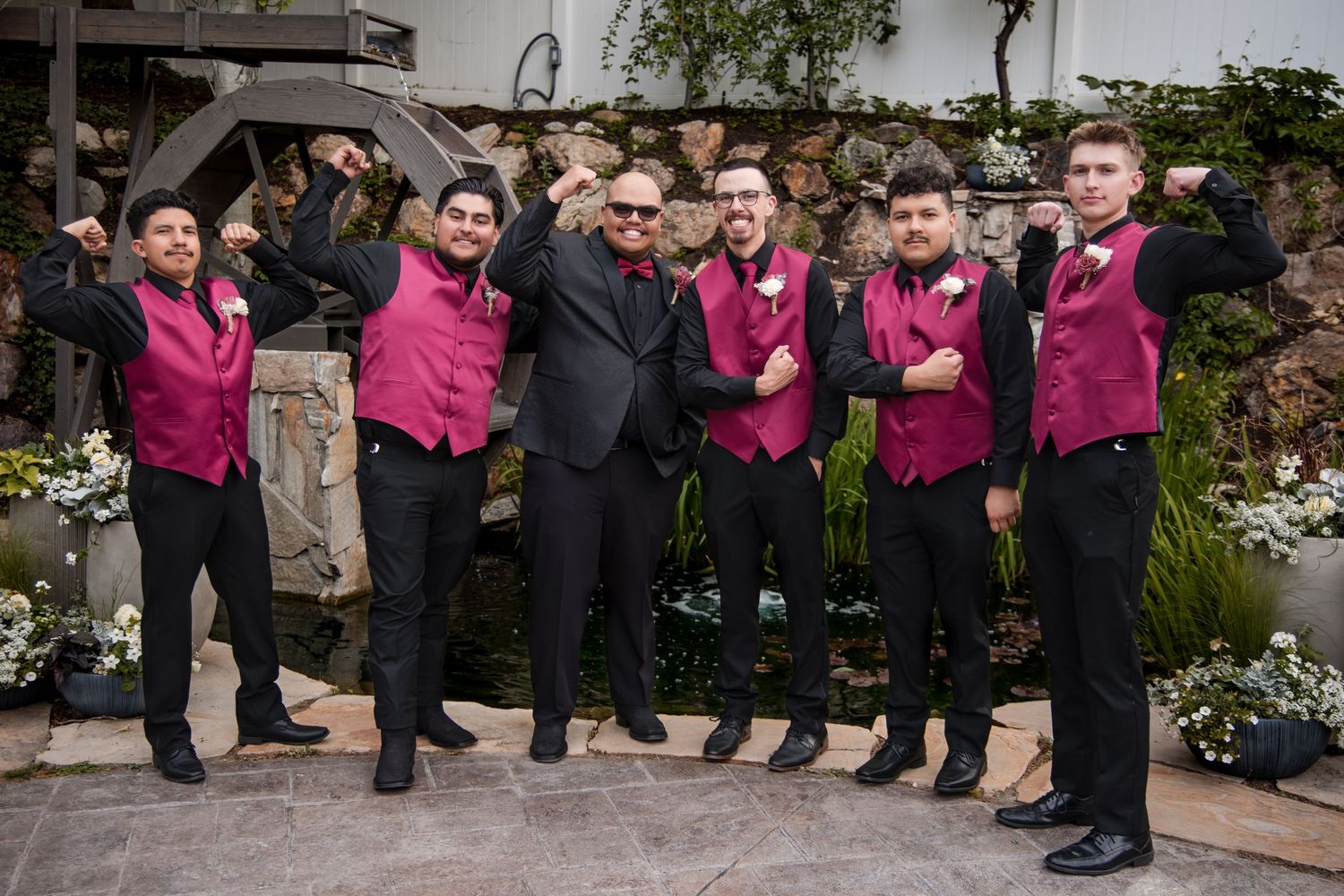 Wedding party members stand in a row outdoors wearing formal attire with pink bridesmaid dresses and dark suits.