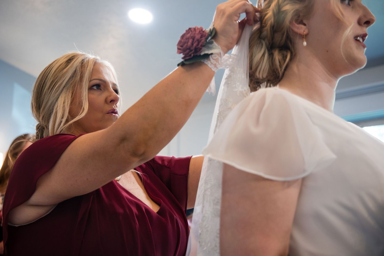 Someone in a burgundy dress helps adjust a white garment in what appears to be a dressing room setting.
