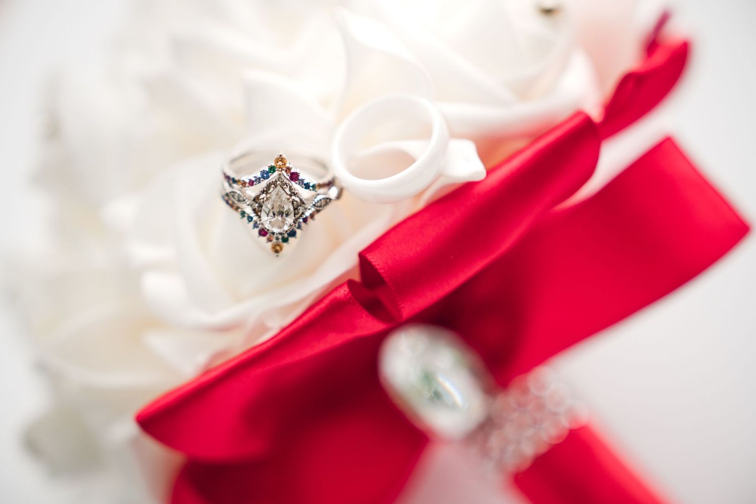 Diamond wedding ring rests against red satin ribbon with white flowers creating an elegant wedding photo composition.