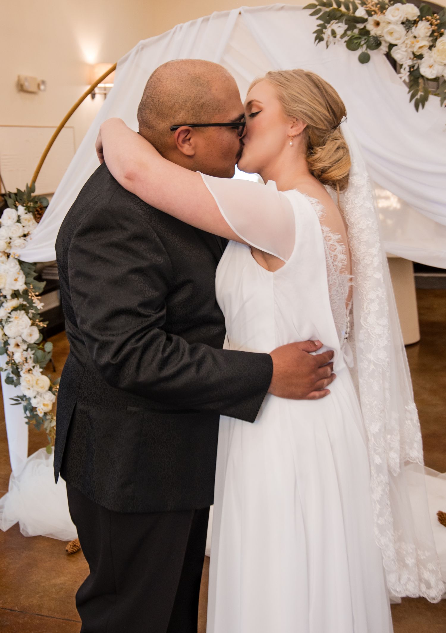 A tender romantic moment at a wedding ceremony under an archway decorated with white flowers and drapery.