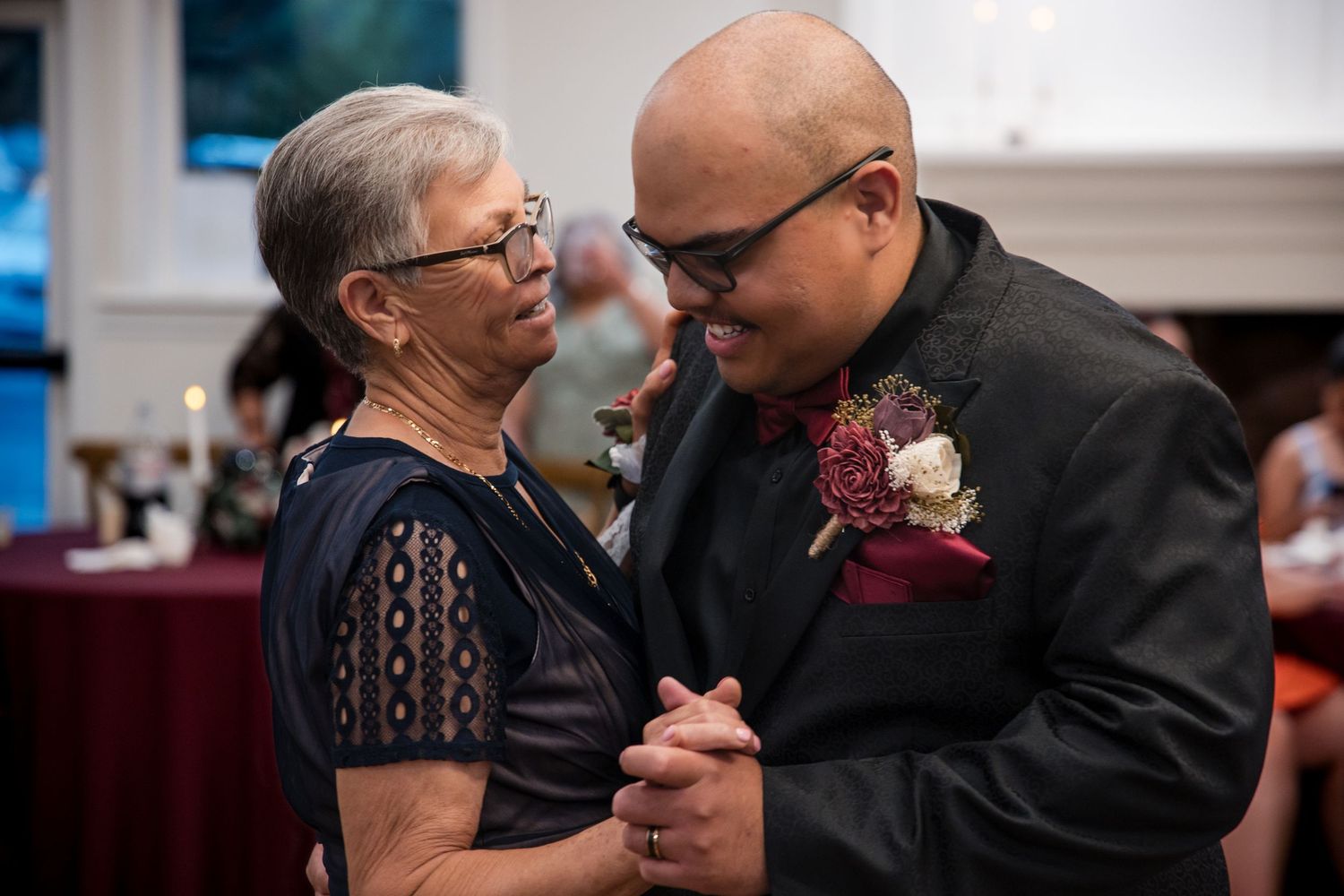 An older woman in a black lace dress dances with a bald man wearing a dark suit with a pink boutonniere at an event.