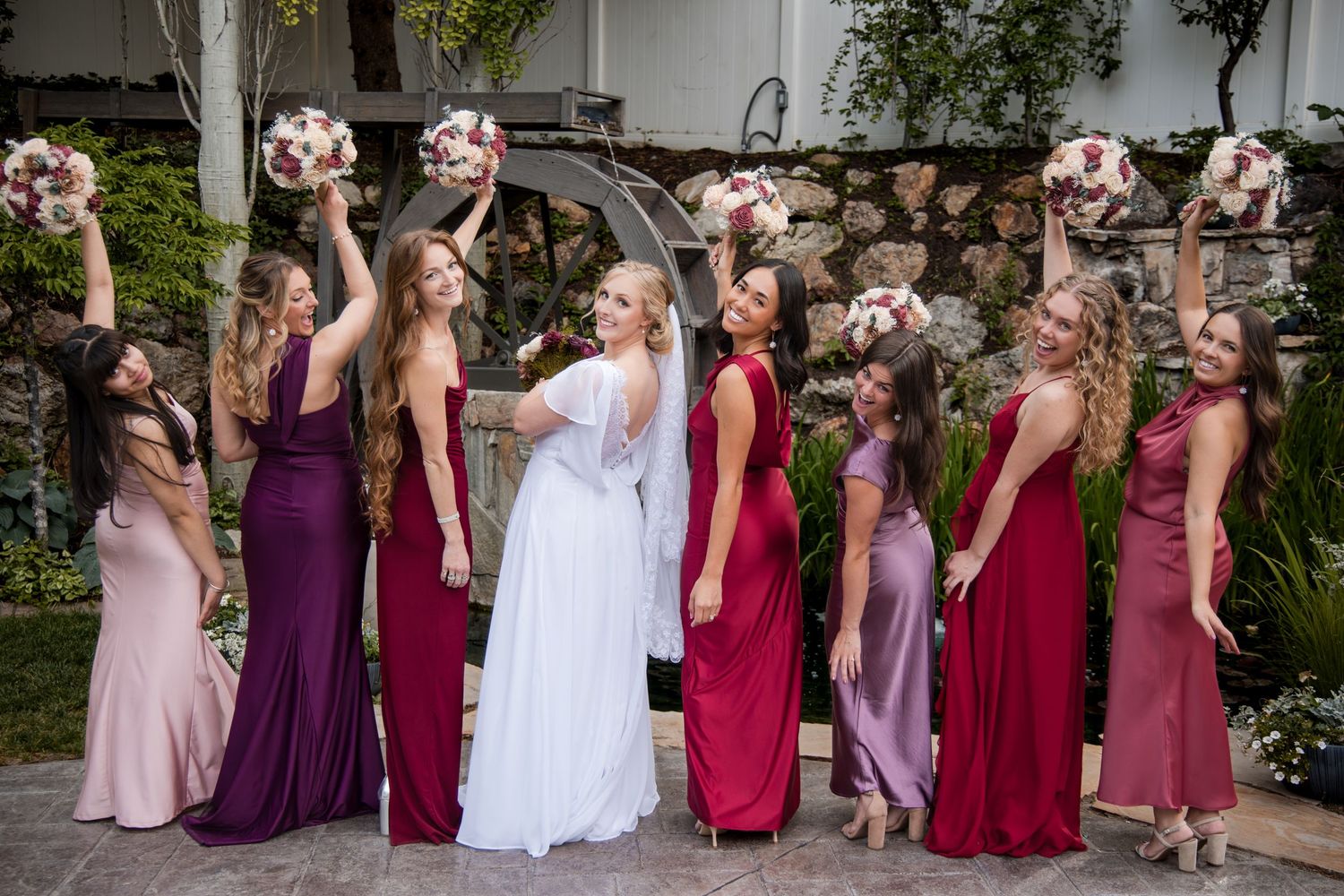 Wedding party standing in line wearing assorted red and purple dresses holding pink floral bouquets in garden setting.