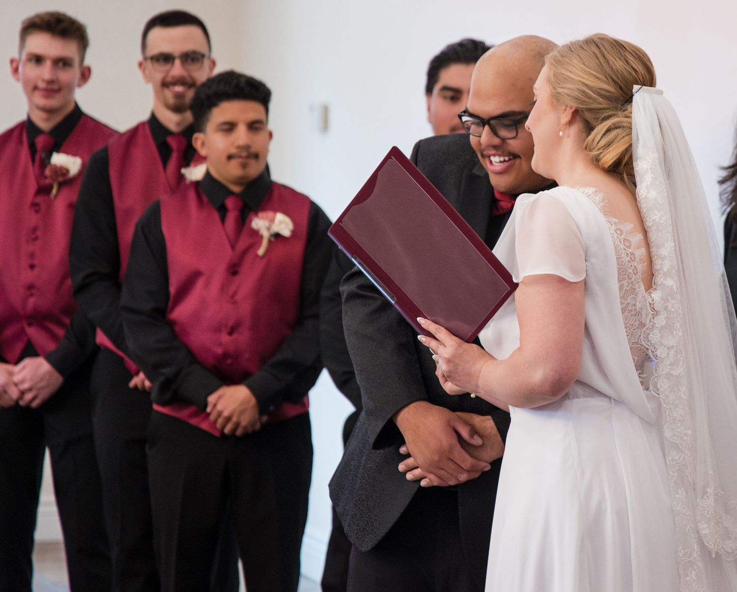 Wedding party in burgundy and black attire stand in a line during ceremony reading from booklets.