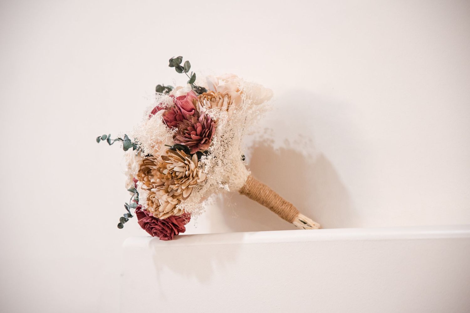 A dried floral bouquet with red and beige flowers cast a soft shadow against a white wall.