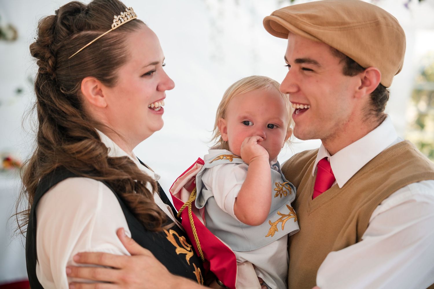 A family of three in vintage-inspired attire smiles together at a wedding while their baby sucks their thumb.