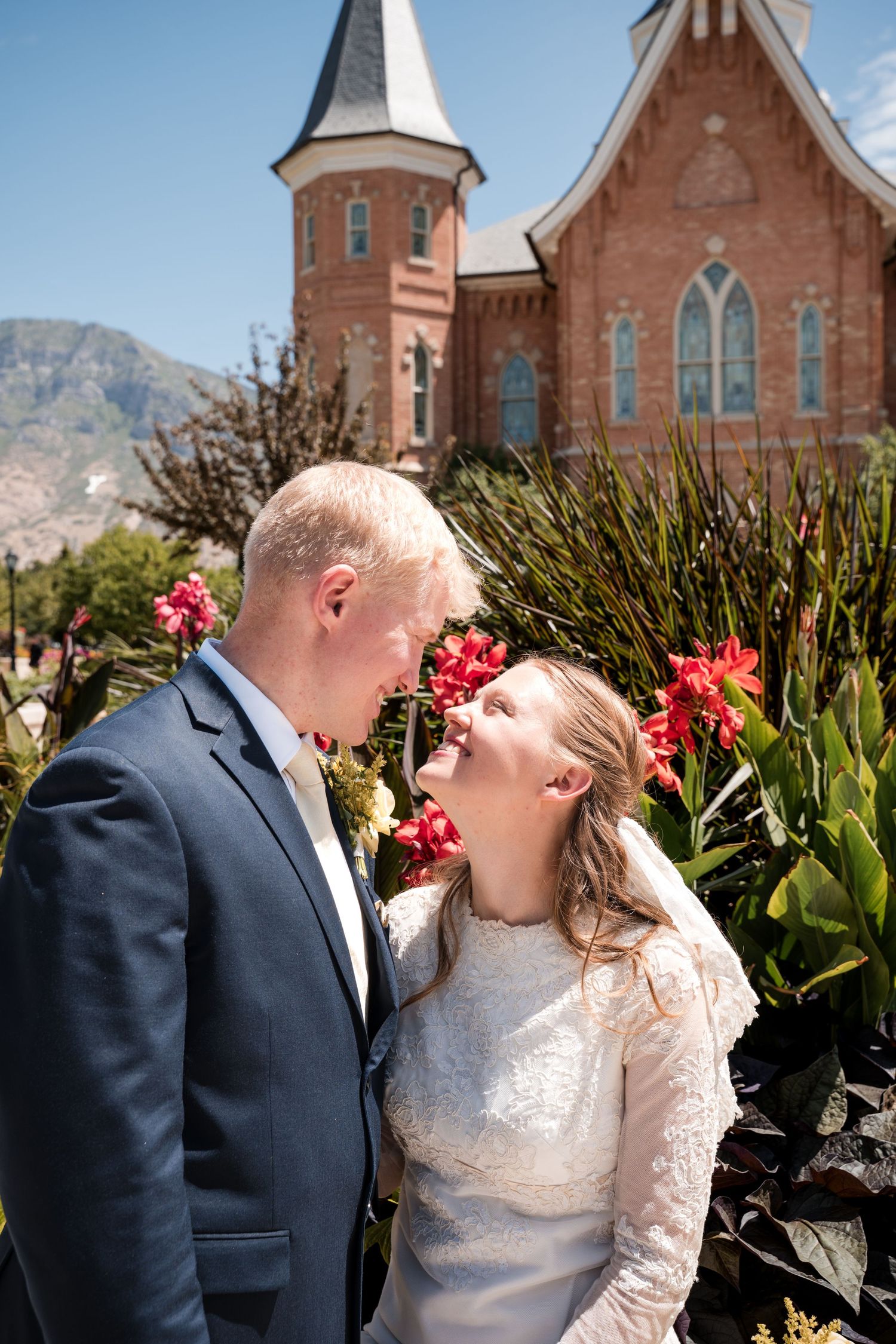A couple shares an intimate moment in front of the Provo City Center LDS temple with red flowers and mountains in the background.
