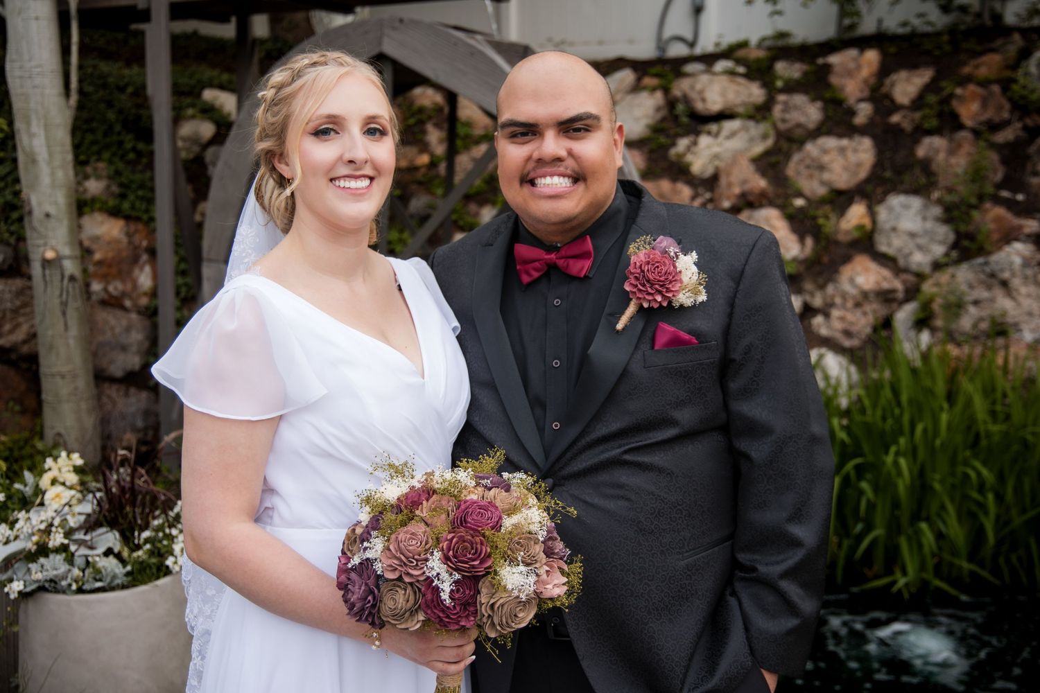 A wedding couple poses together outdoors, with the bride in white dress holding a burgundy bouquet and groom in dark suit.