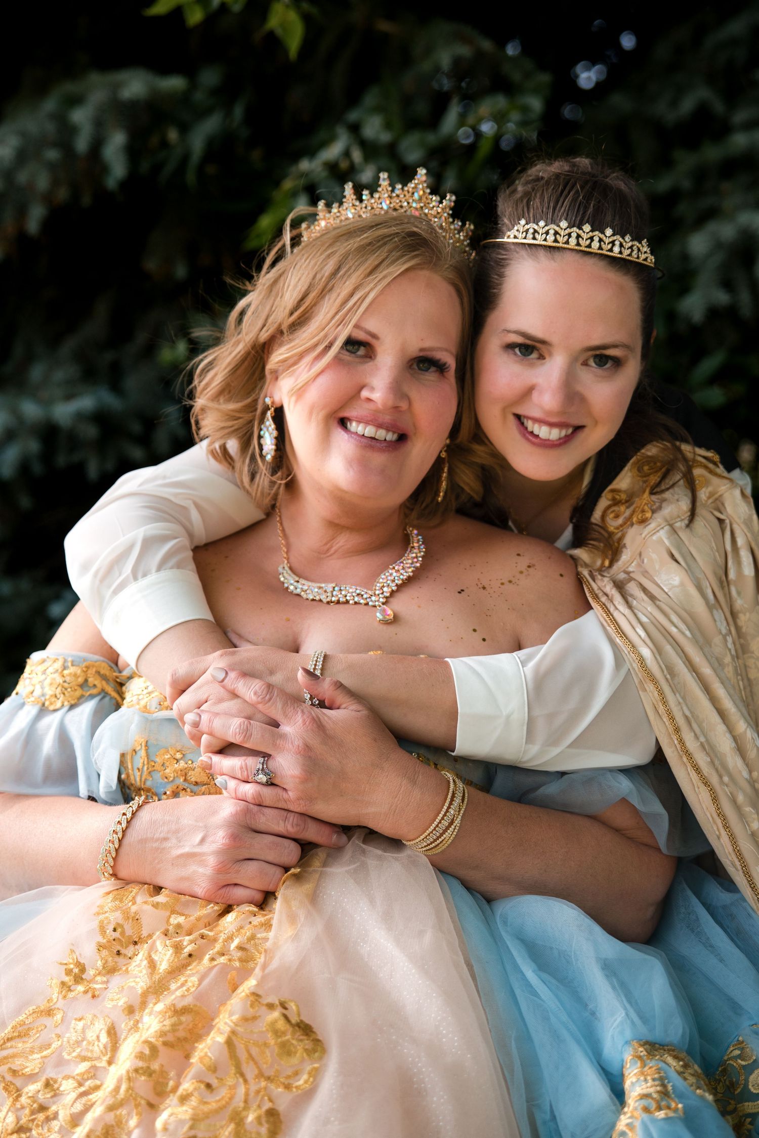 Two people in elegant gowns with tiaras and jewelry embrace while posing together outdoors against a dark foliage background.