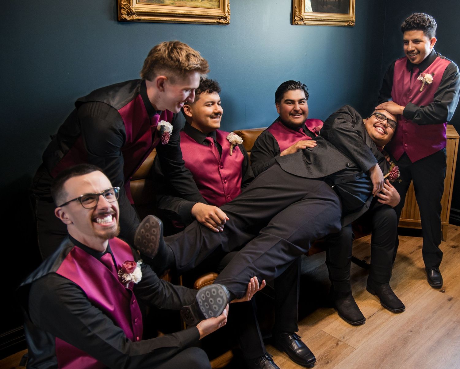 A group of smiling wedding party members in burgundy suits and vests relax together on a blue couch.