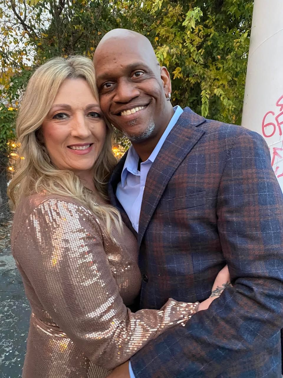 A couple dressed in a sparkly bronze dress and plaid blazer pose together warmly outdoors with autumn foliage in background.