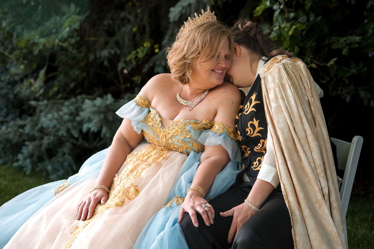 A romantic fairytale couple in elaborate period costumes share an intimate moment on a garden bench.