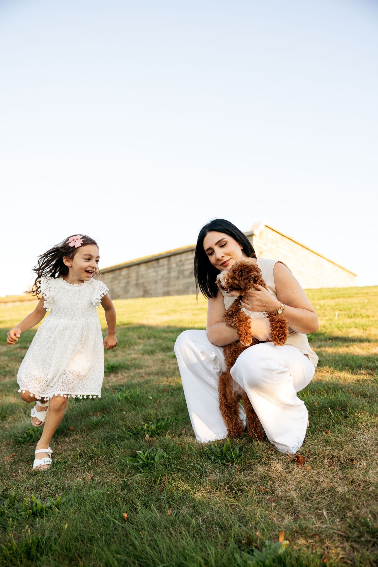 Two people in white dresses laugh and roll together on a grassy hillside at sunset.