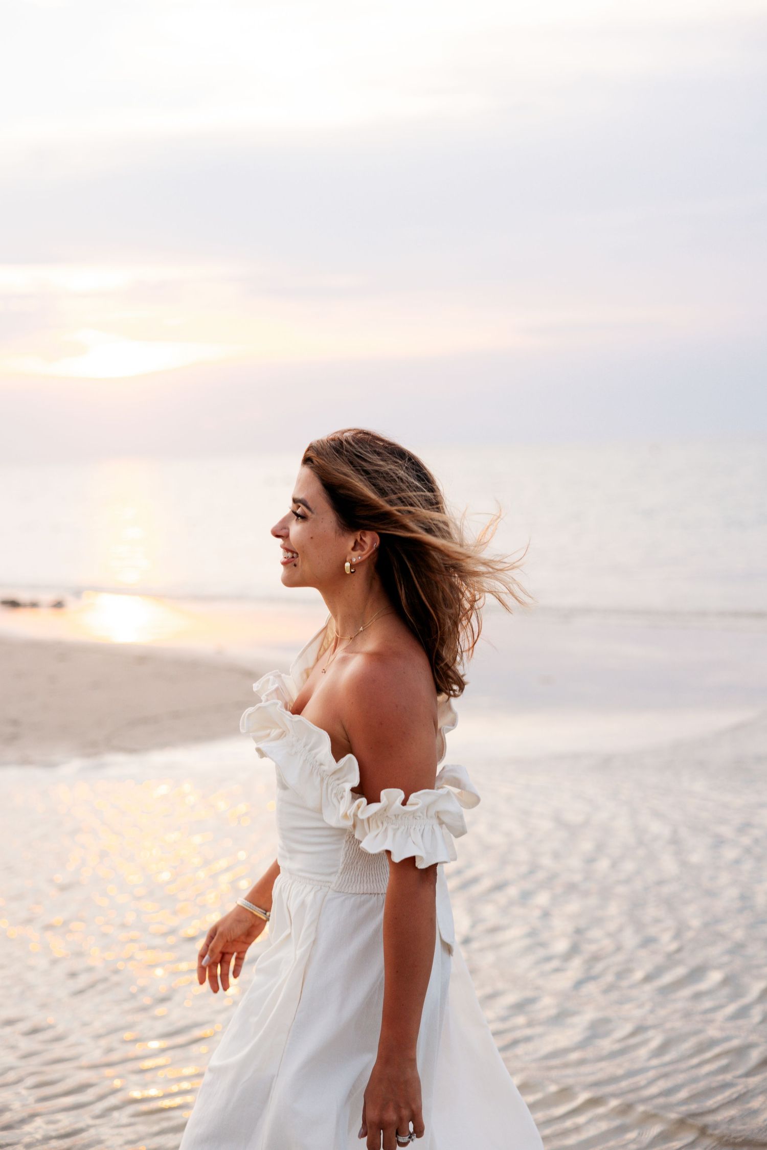A person in a white off-shoulder wedding dress stands on a sandy beach at sunset with wind-blown hair.