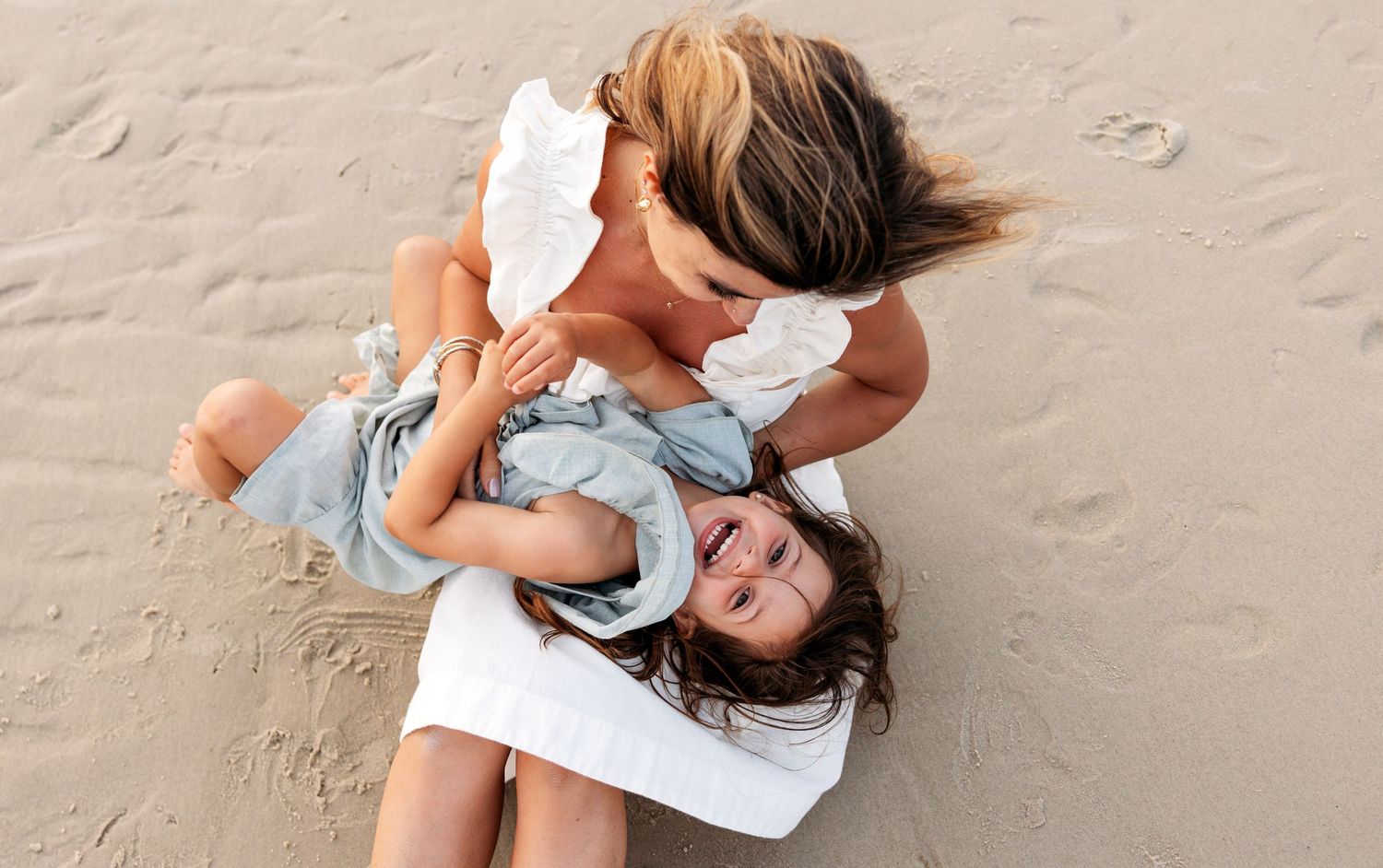 People wearing white and blue clothing lay on sandy beach during tender moment of laughter and connection.