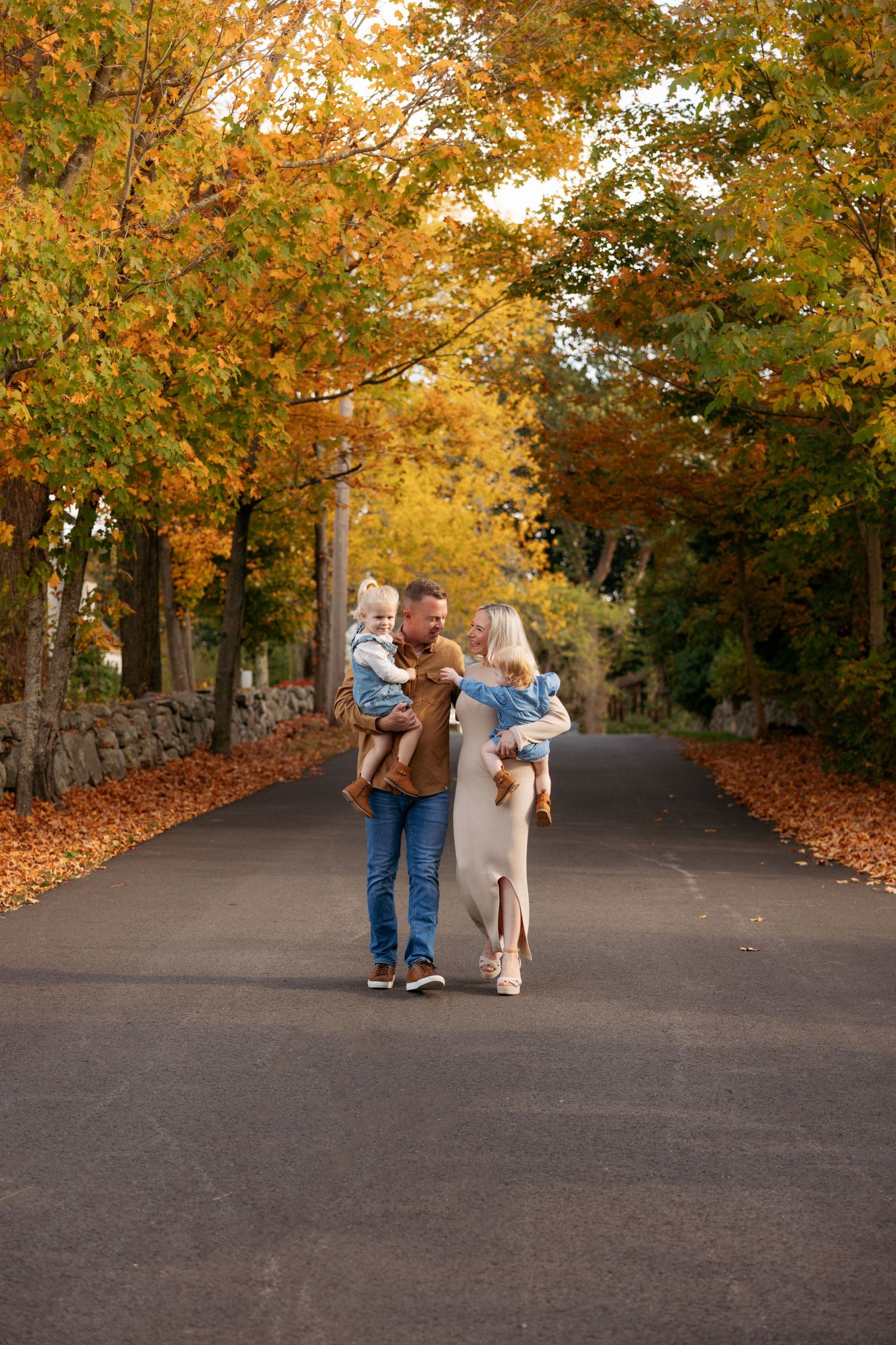 A couple walks together on a tree-lined autumn path covered in fallen orange leaves.