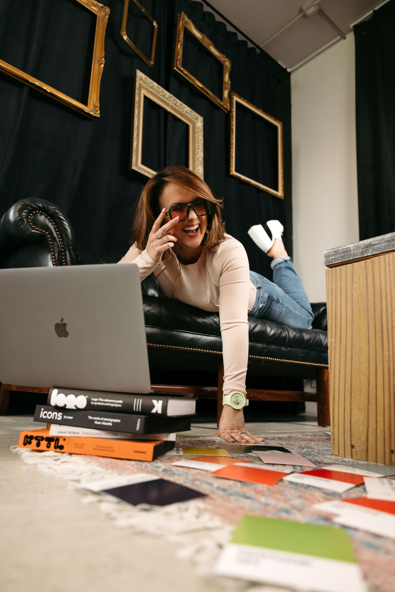 Woman laughing at laptop on floor surrounded by design books and color swatches, lying by black leather sofa with gold frames.